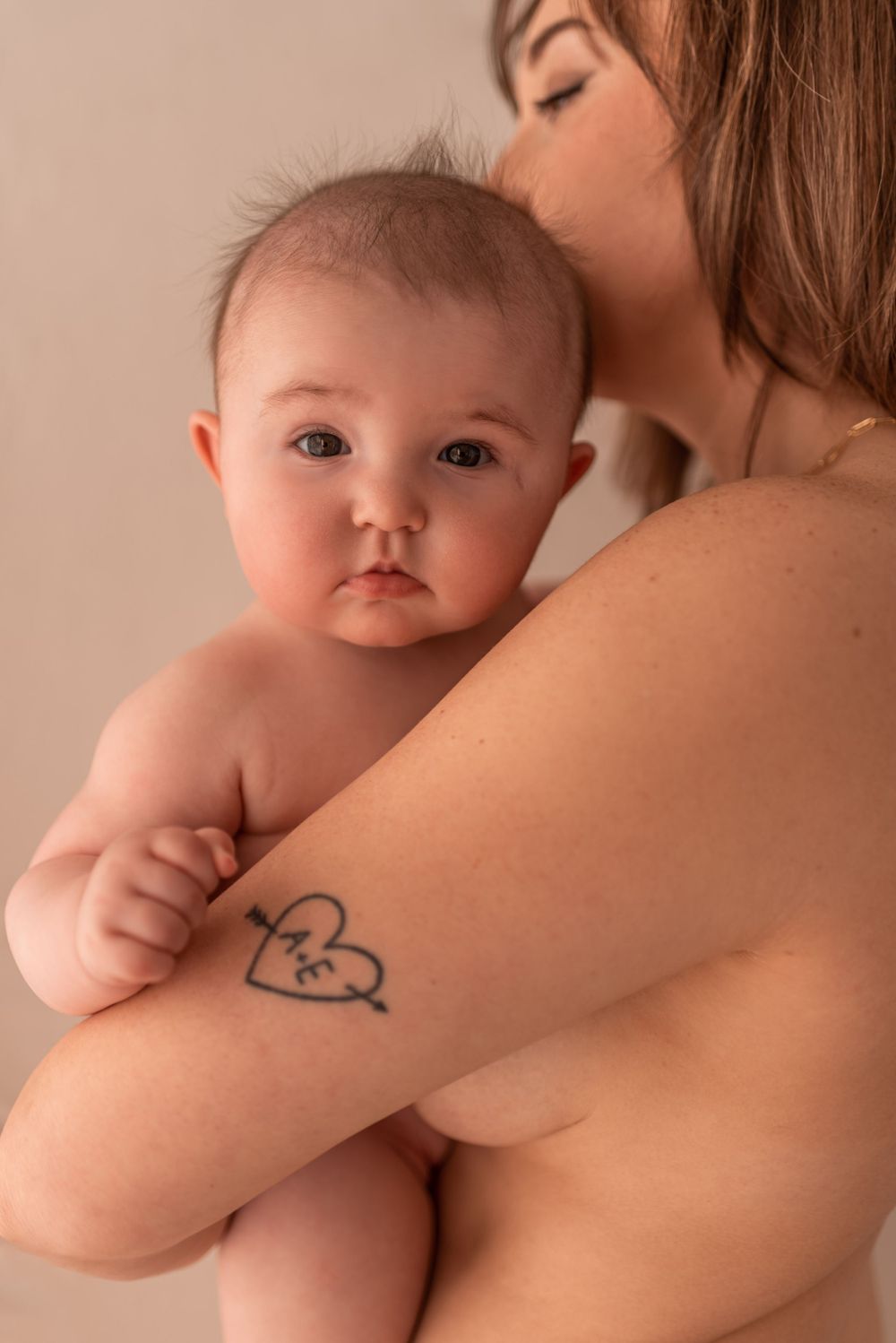Artistic portrait showing mother with heart tattoo holding infant against neutral background.