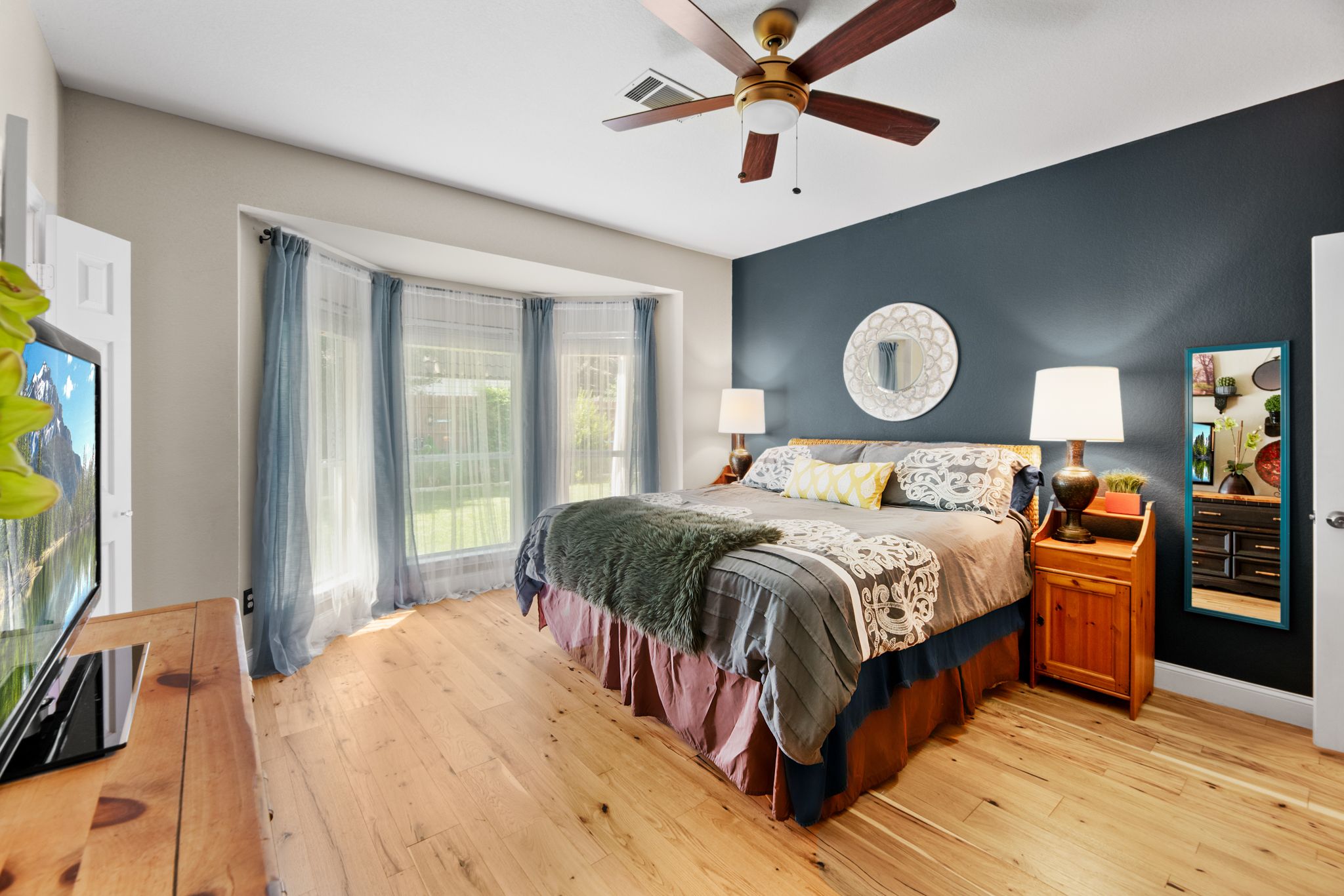 Spacious master bedroom with navy accent wall, large bay windows, wooden floors and ceiling fan.