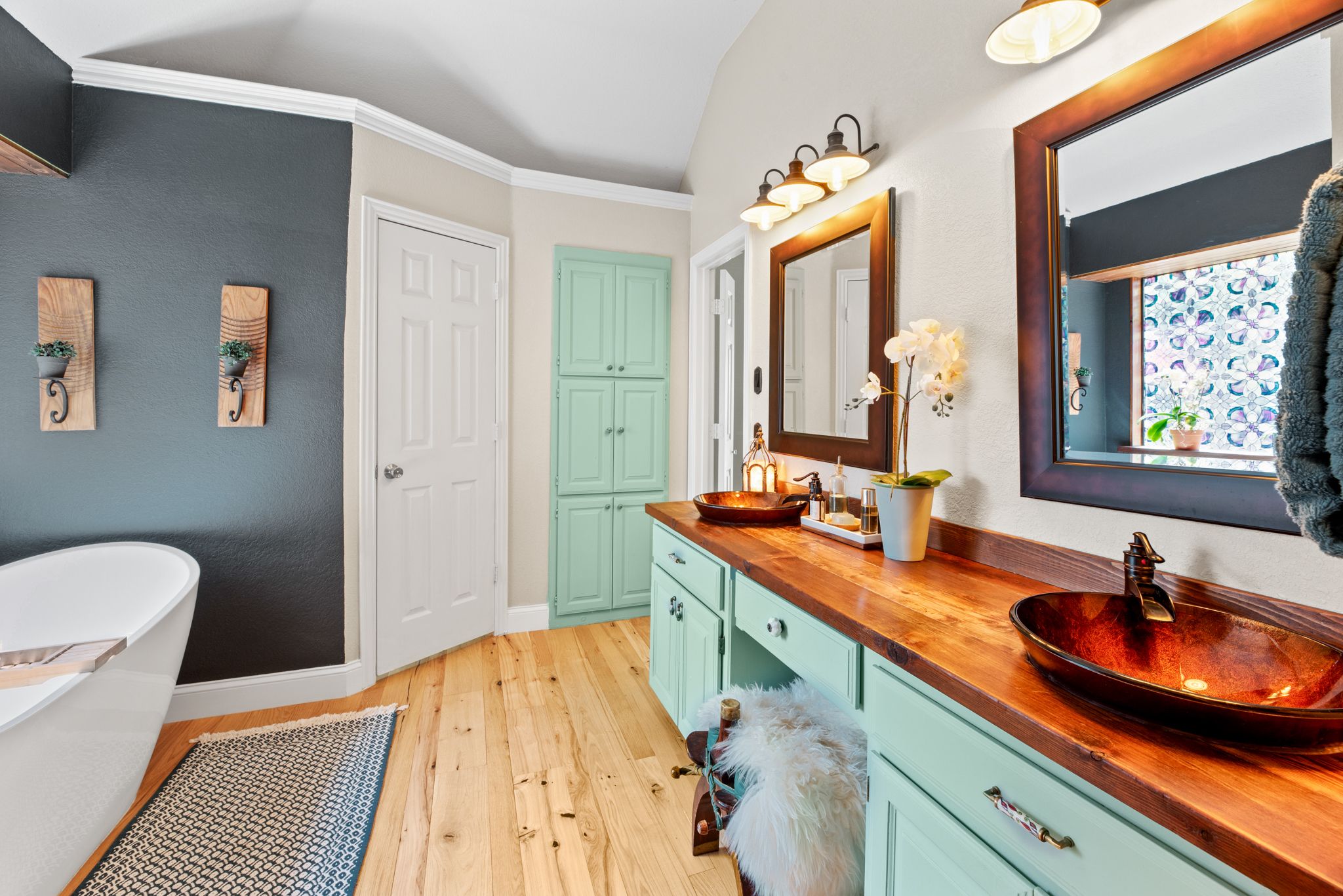 Modern farmhouse bathroom features mint cabinets, wood countertops, copper vessel sink and freestanding tub against dark accent wall.