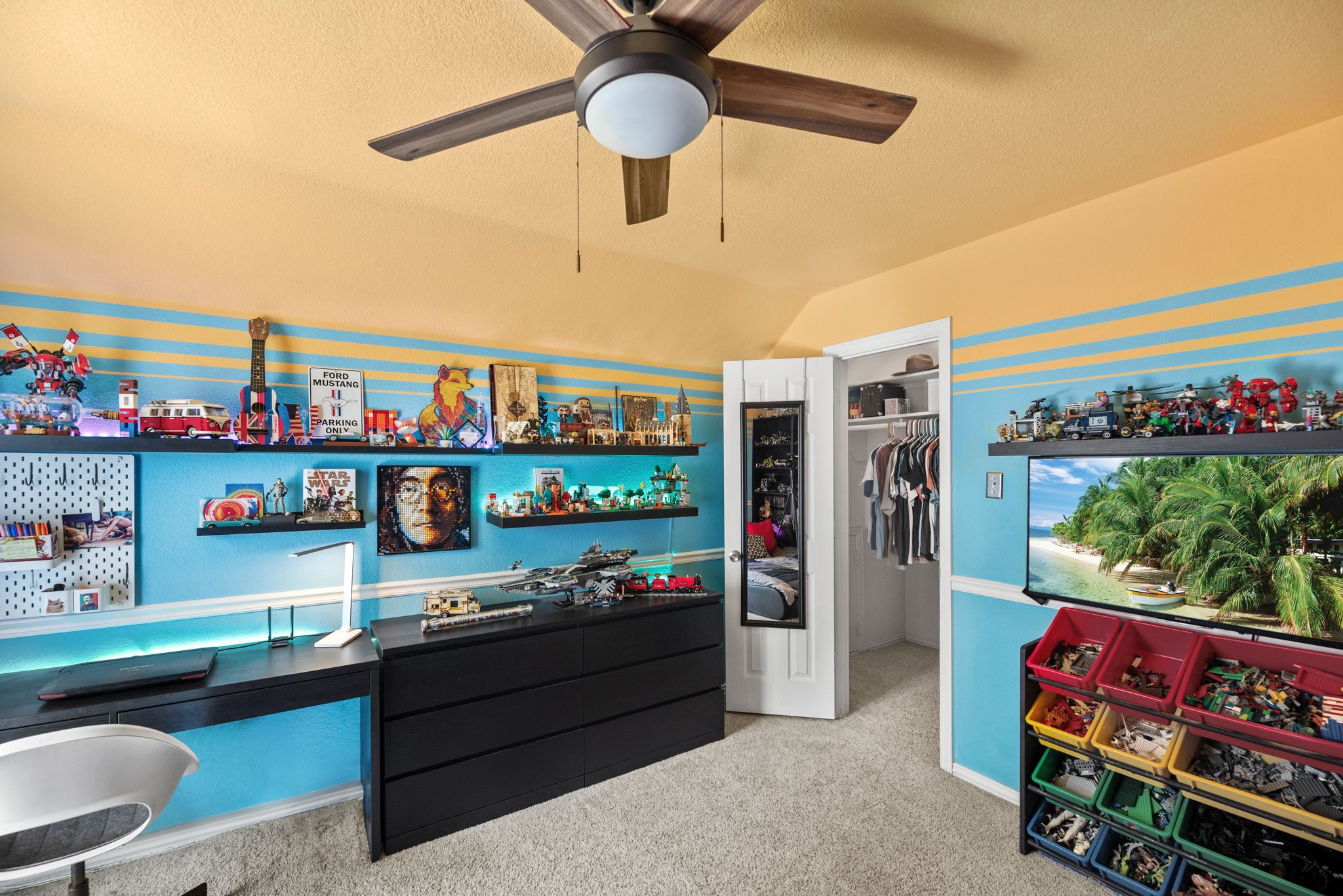 Modern gaming room with multiple monitors, gaming consoles, and storage shelves against turquoise blue striped walls.