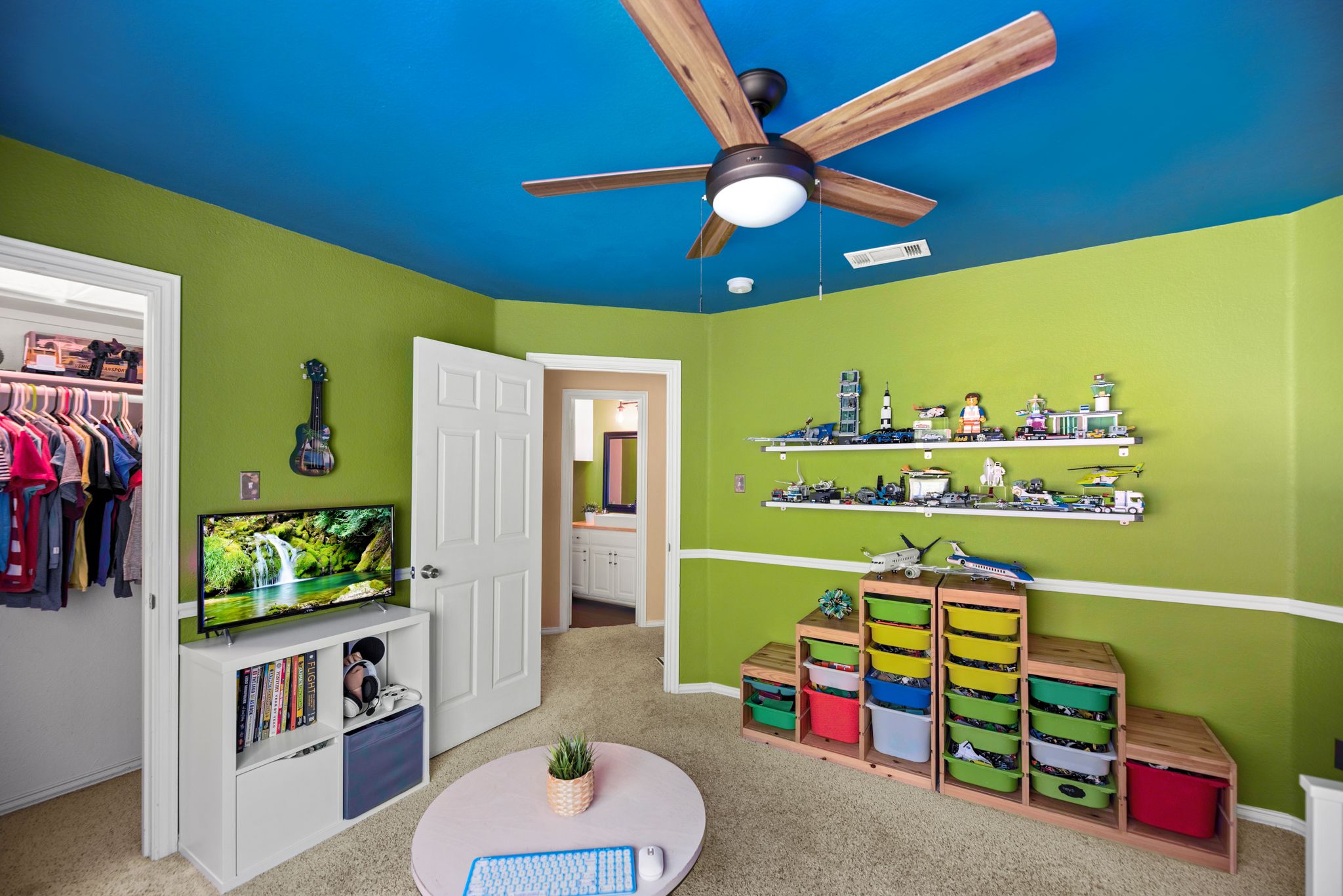 A colorful children's playroom with bright green walls, floating shelves, storage bins, and a modern ceiling fan.