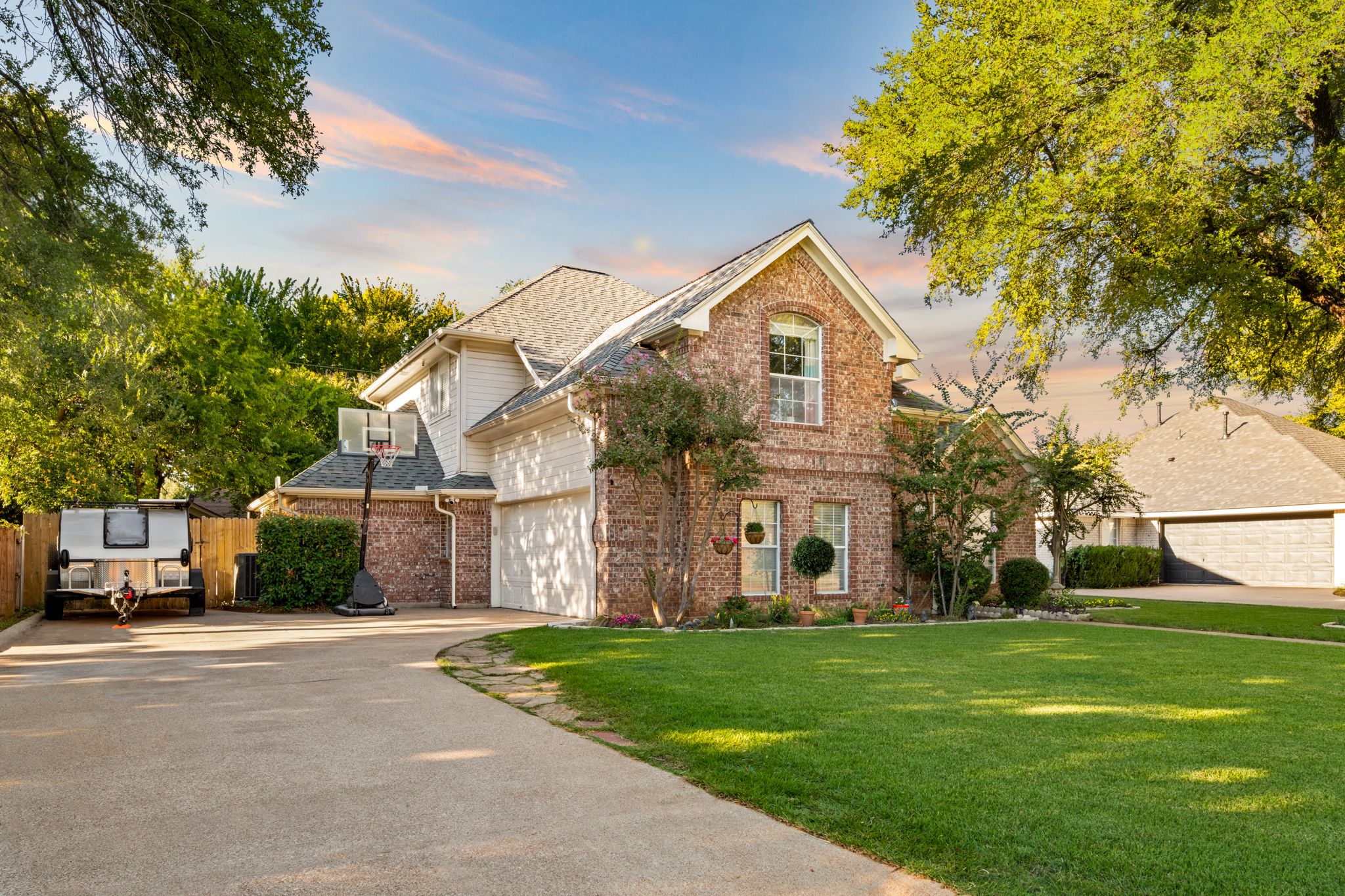 Exterior view of a modern brick house with green lawn and mature trees in a residential neighborhood at sunset.