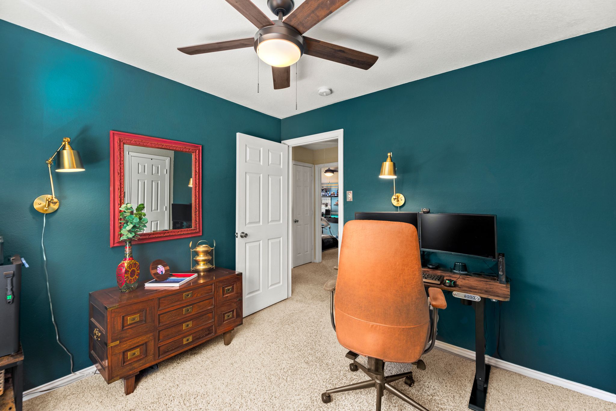 A home office with dark teal walls, a wooden dresser, orange chair, red framed mirror, and brass sconces.