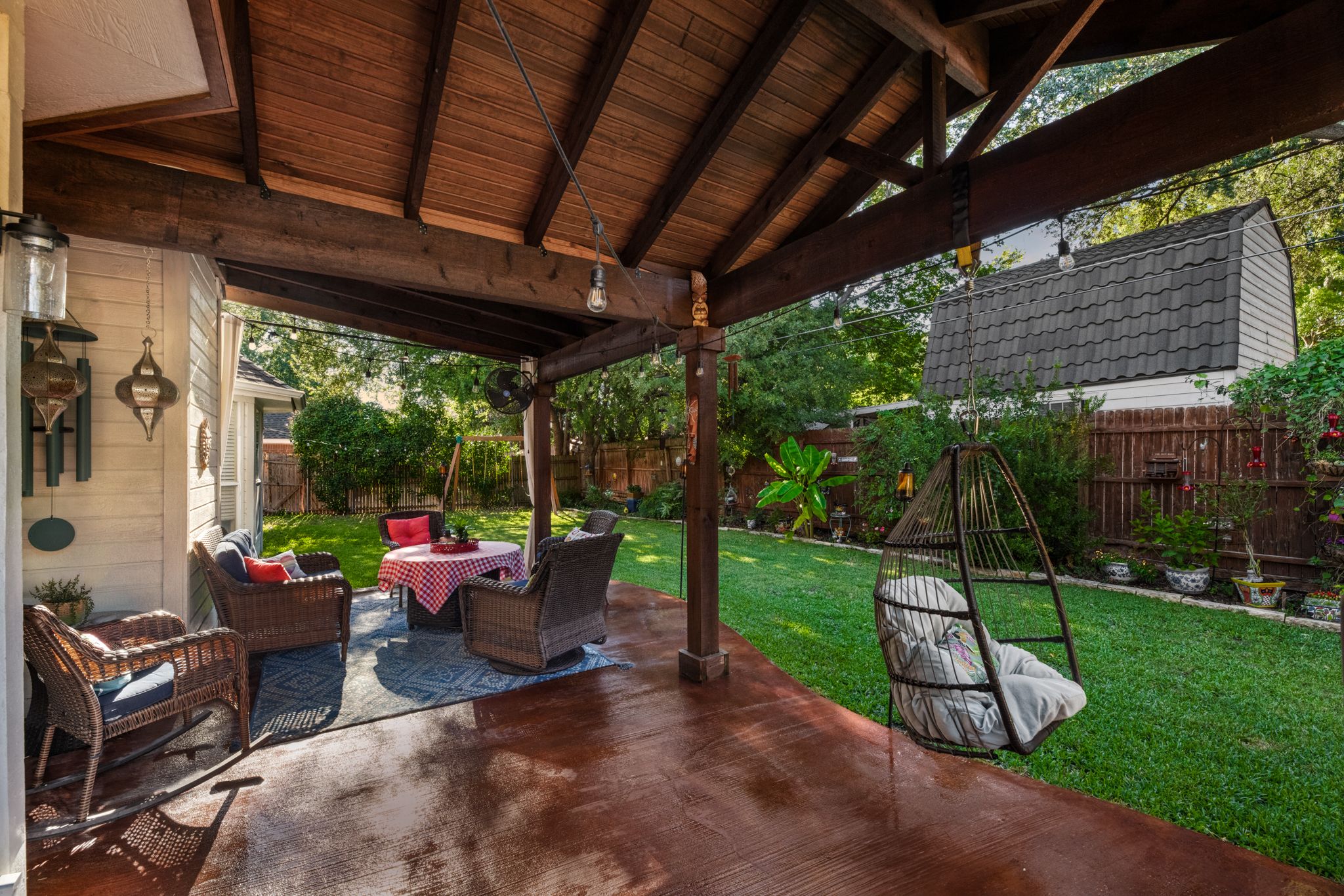 Cozy covered patio with outdoor furniture overlooks a green backyard with swing set under a wooden pergola.