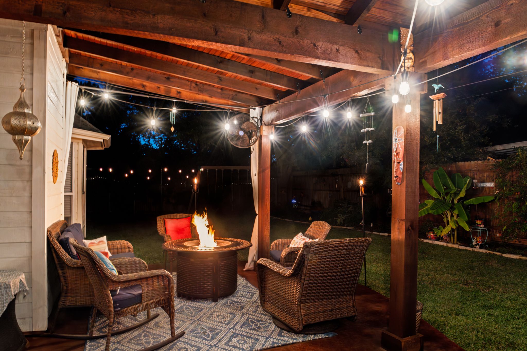 Cozy outdoor patio with wicker chairs around a fire pit under wooden pergola with string lights creating warm ambiance.