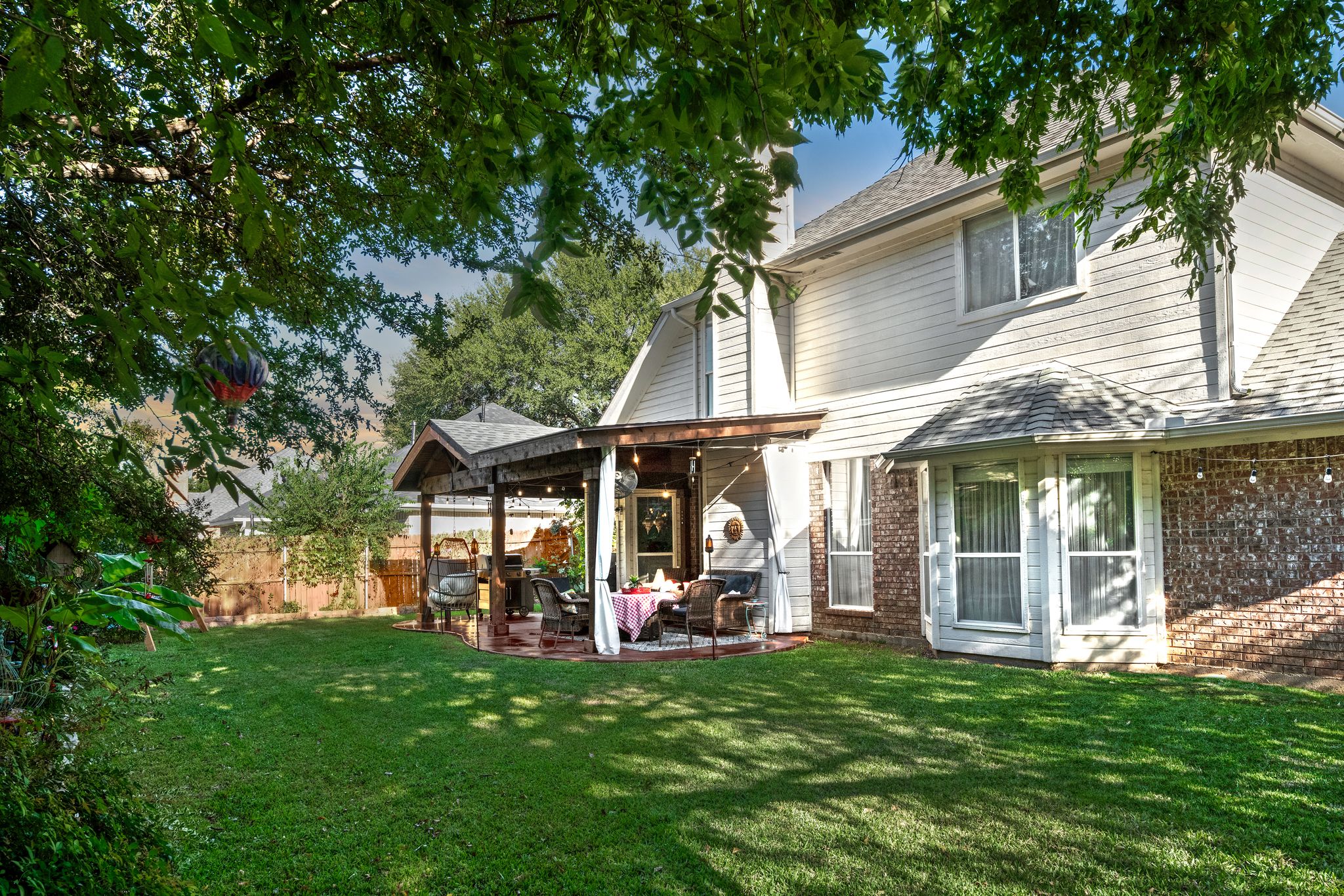 Beautiful two-story white house with covered patio and well-maintained backyard lawn surrounded by lush trees.