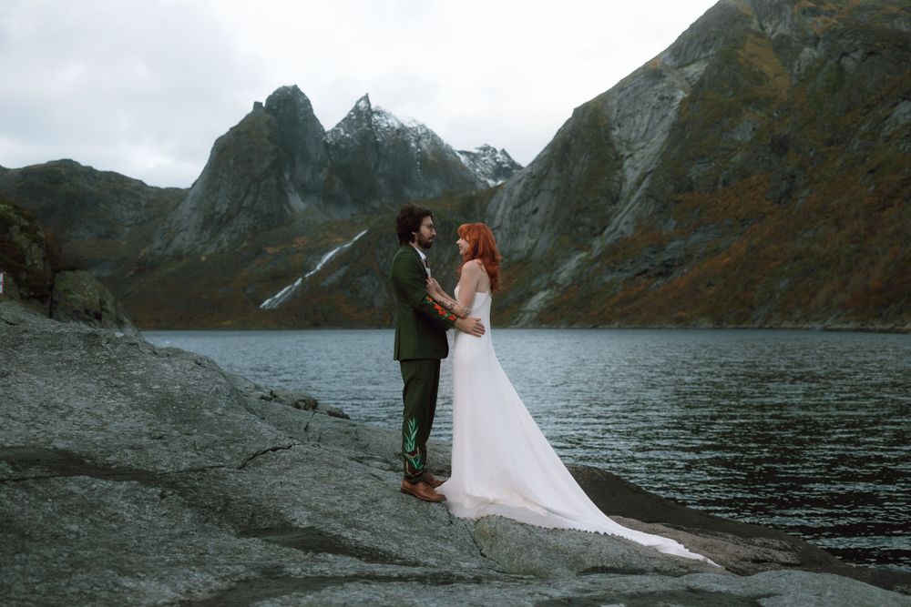 Wedding couple stands together on rocky shore with dramatic mountain lake landscape.
