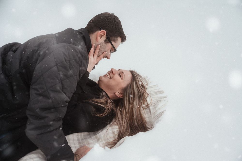 Romantic couple lying in snow during winter photoshoot with falling snowflakes.