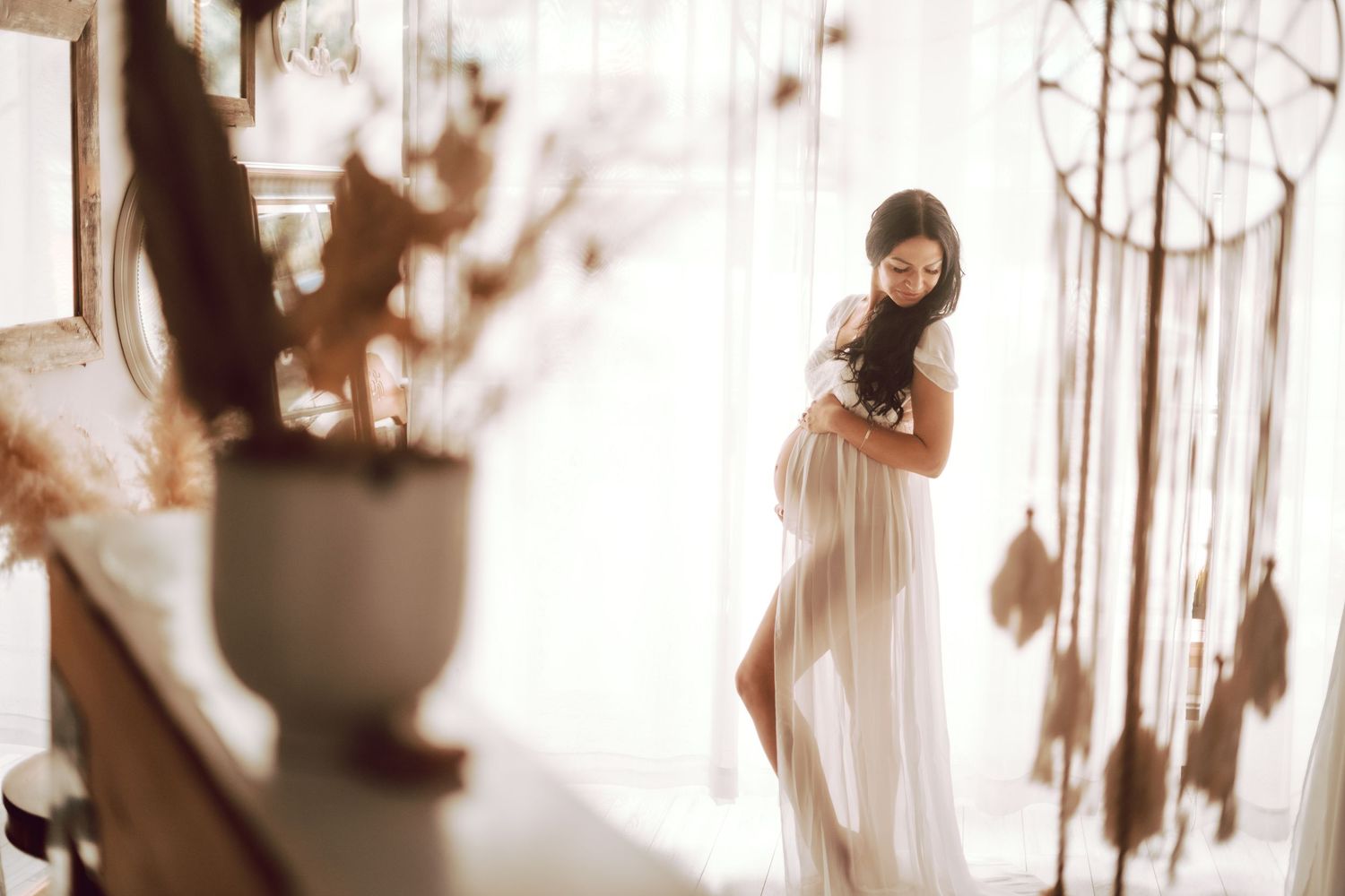 Dreamy maternity photo in white flowing dress with soft natural lighting through sheer curtains and decorative potted plants.