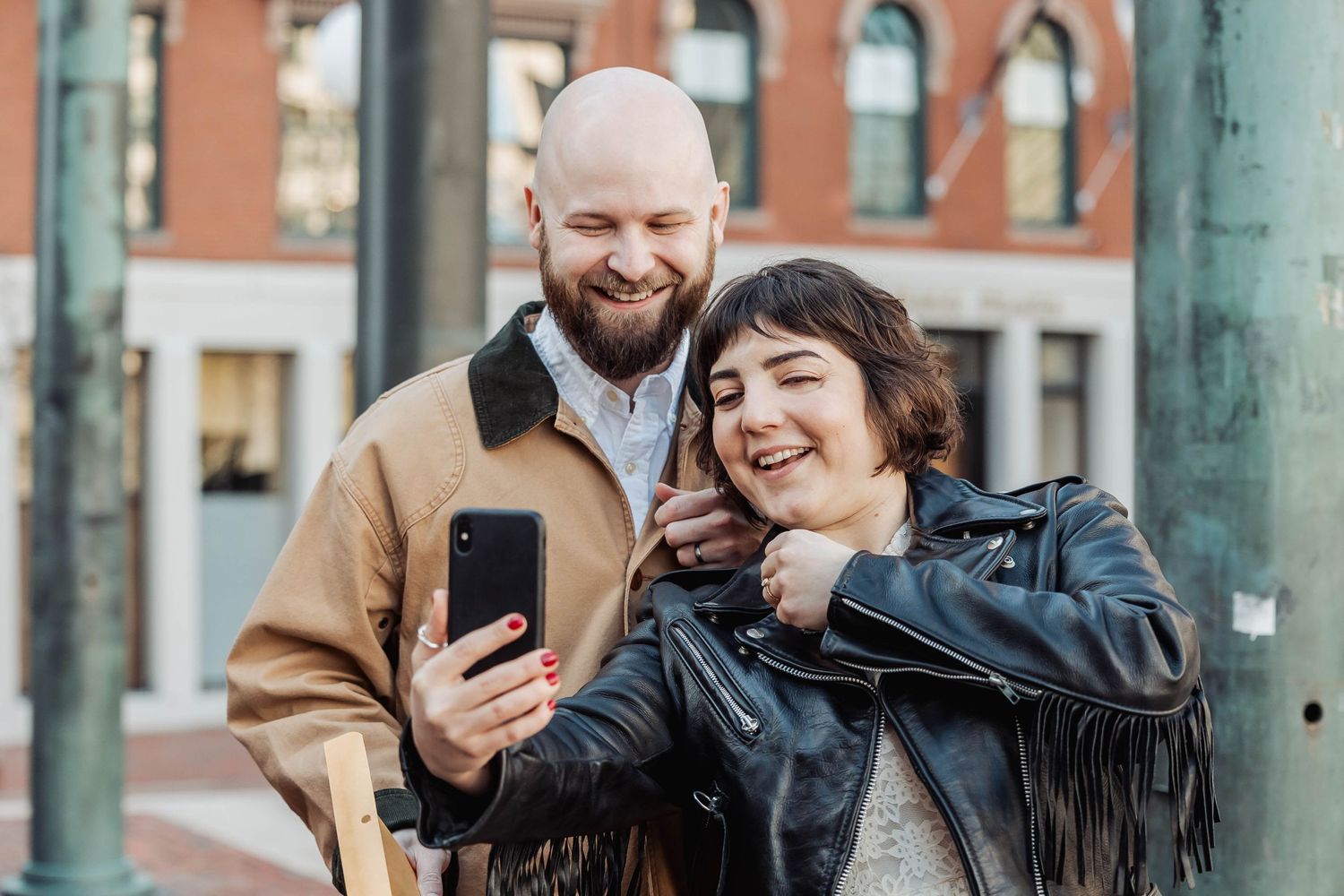 A couple laughs while taking selfies together on an urban street.