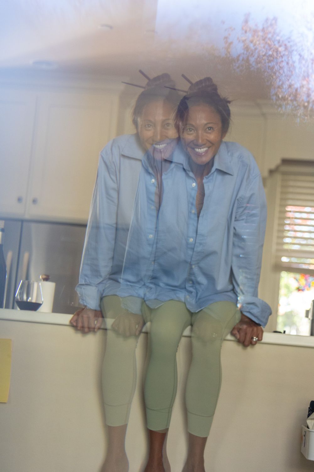 Mirror reflection shows woman wearing denim shirt and olive pants sitting casually on counter in indoor setting.