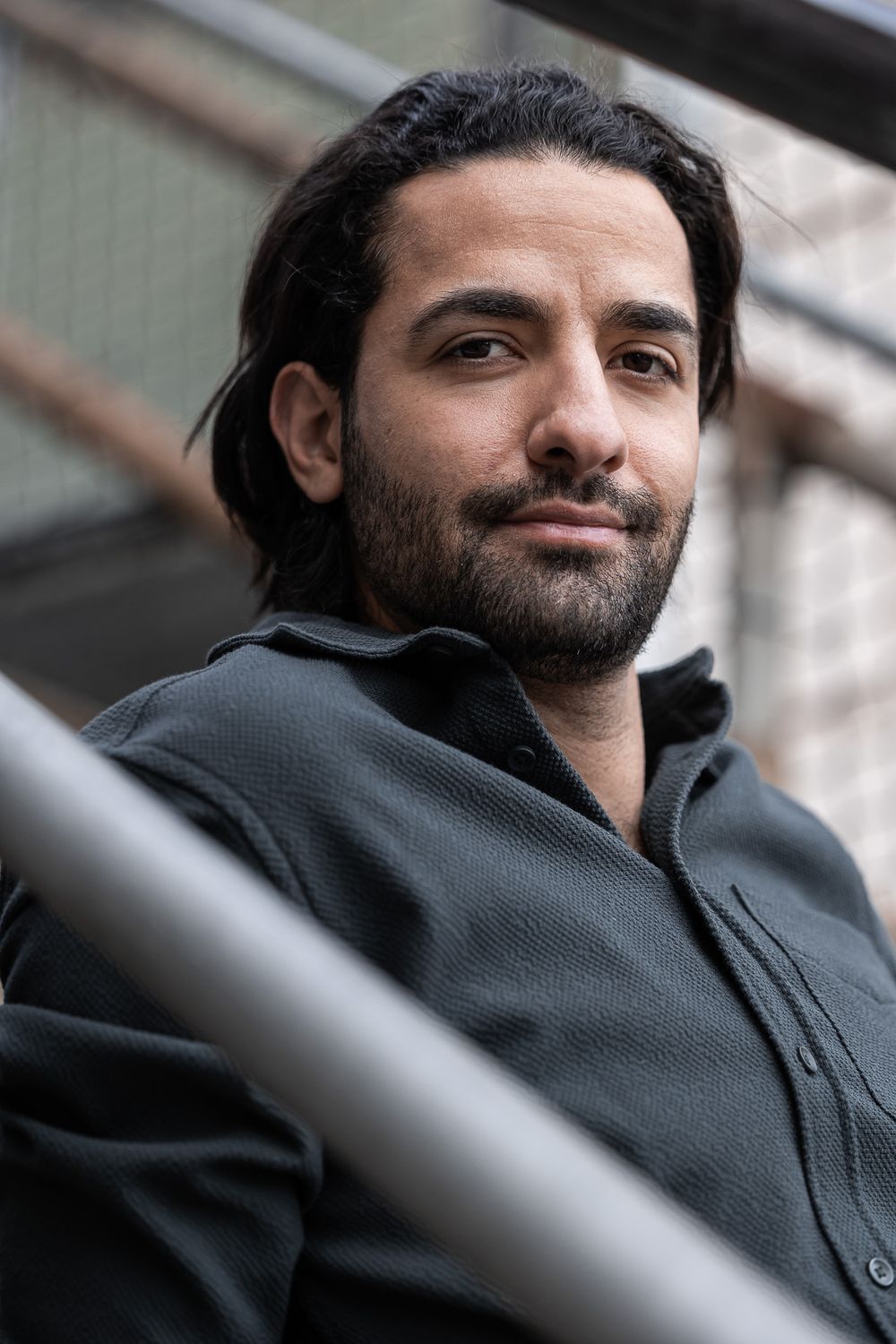 Close-up portrait of a person with dark hair and beard wearing a black zip-up jacket against an outdoor background.