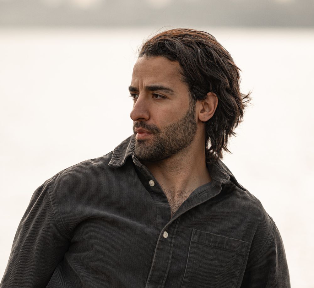 Profile view of a person with dark wavy hair and full beard wearing a black button-up shirt looking off into the distance.
