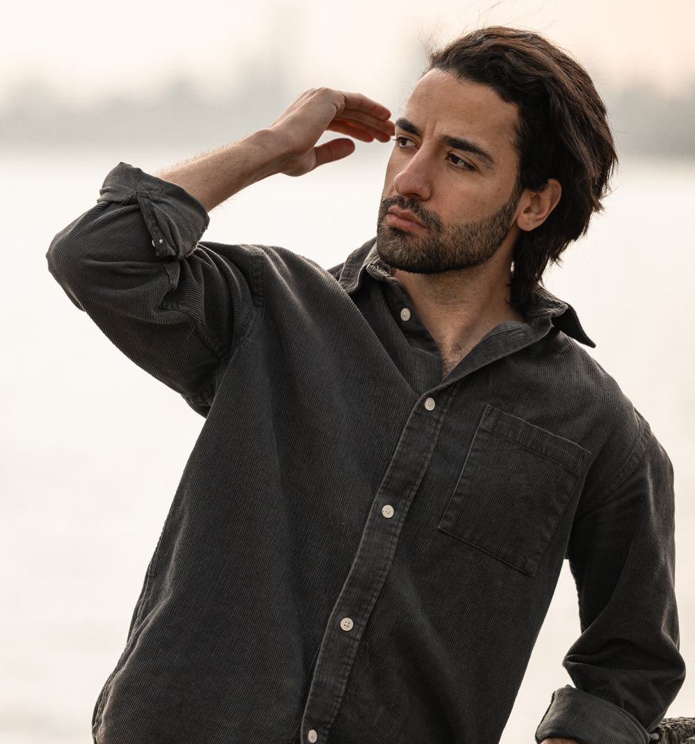 Long-haired man in dark button-up shirt poses outdoors against blurred water background at sunset.
