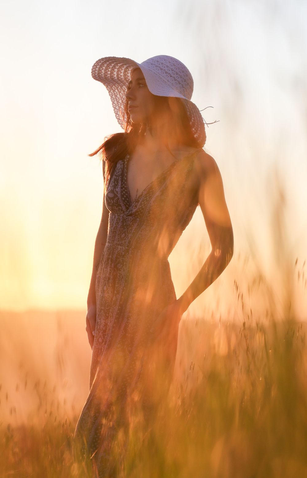 Sunlit figure in white dress and hat standing in golden field at sunset.