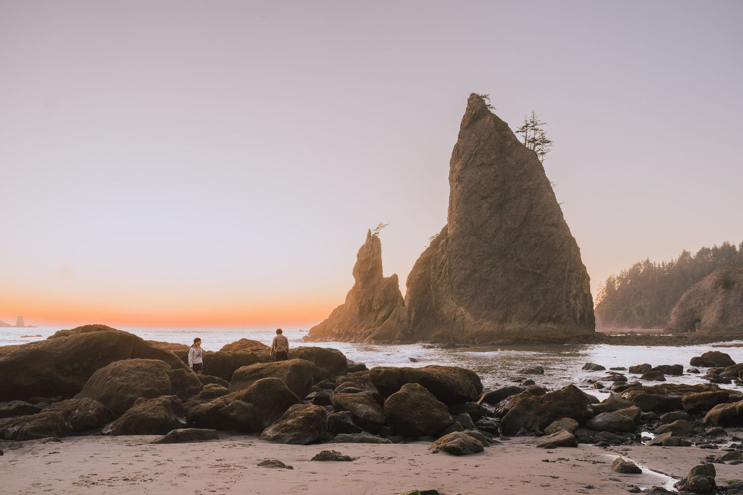 a young couple walking along an Oregon beach within the rocks as the sun sets against the ocean