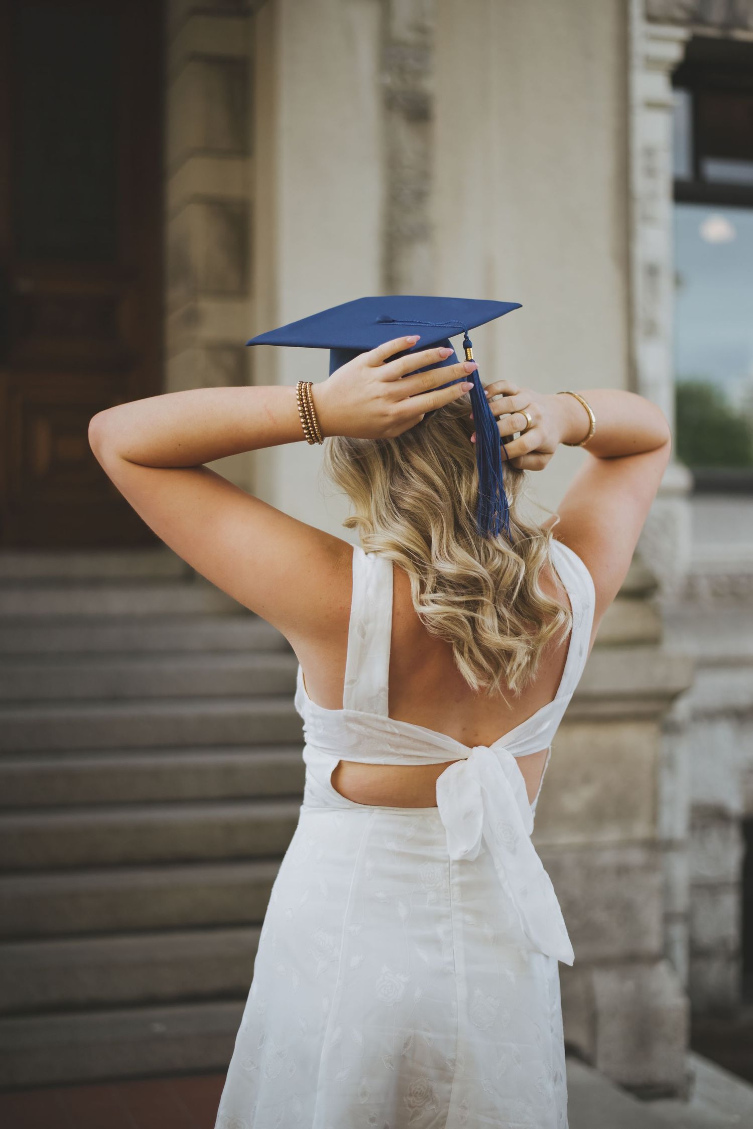 A teenaged girl wearing a white sundress and blue graduation cap adjusts her cap while standing on stone steps on a sunny day.