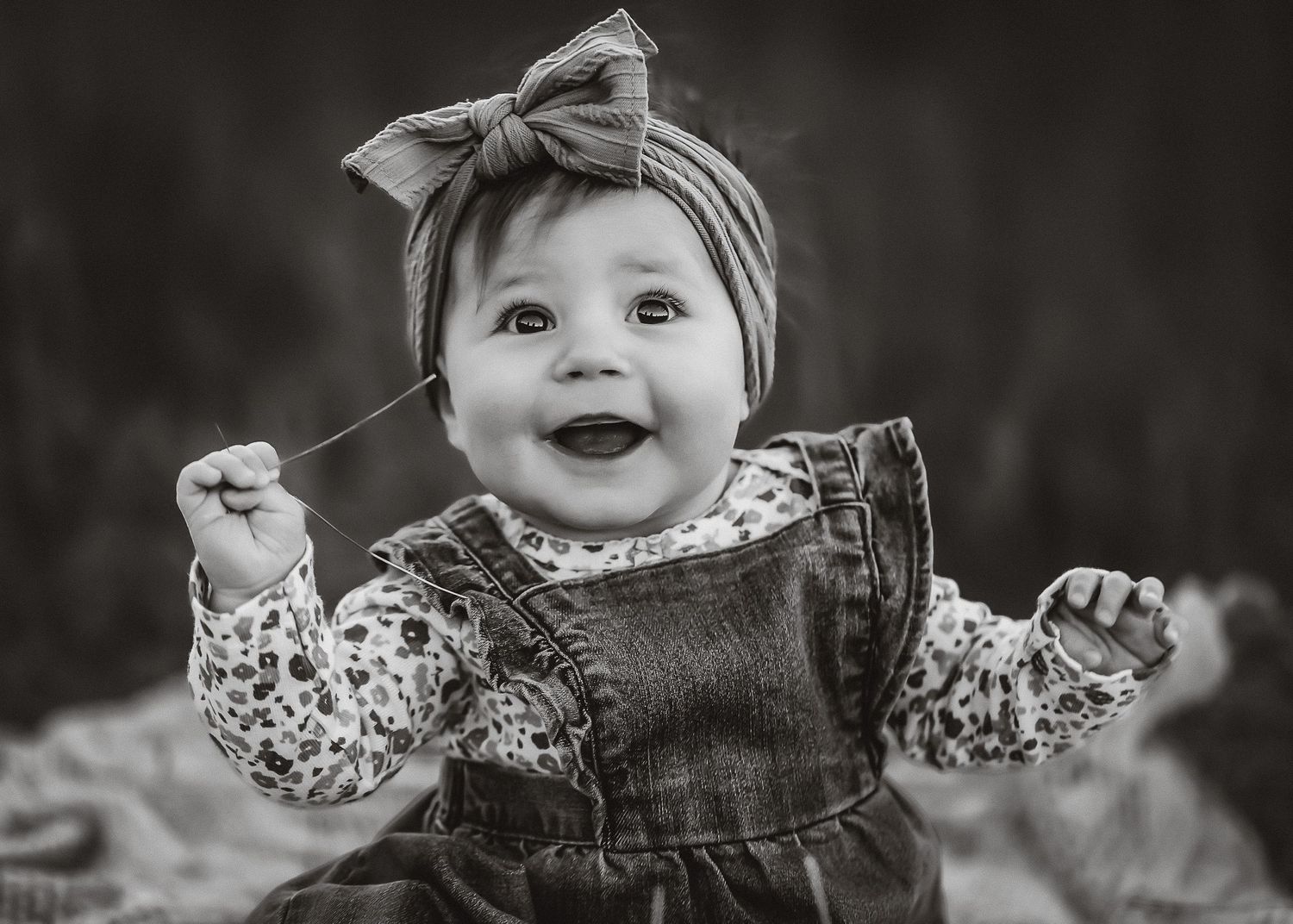 Black and white portrait of baby in denim overalls and bow headband showing pure joy.