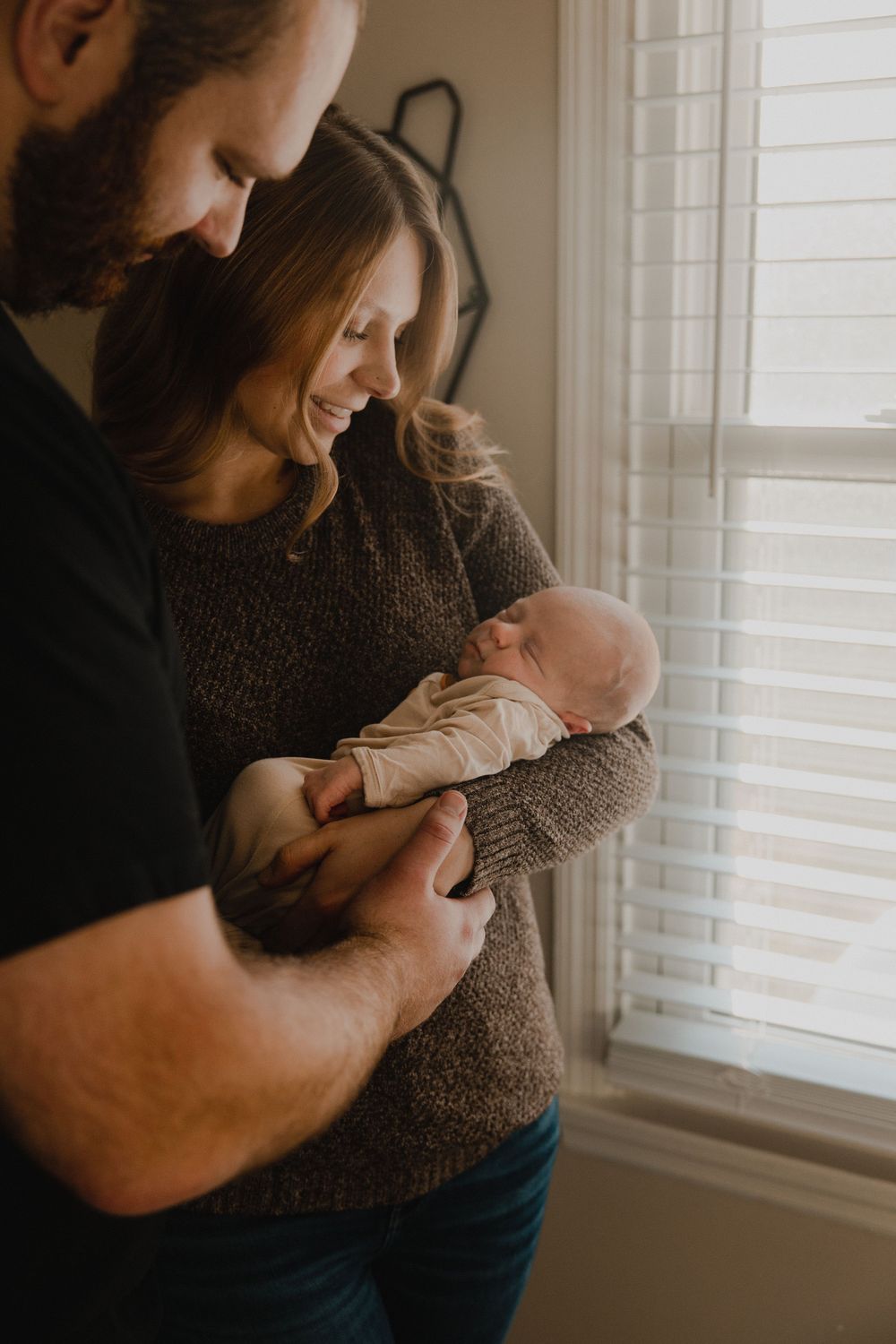 Intimate family portrait by a window showing parents sharing a joyful moment with their sleeping infant.