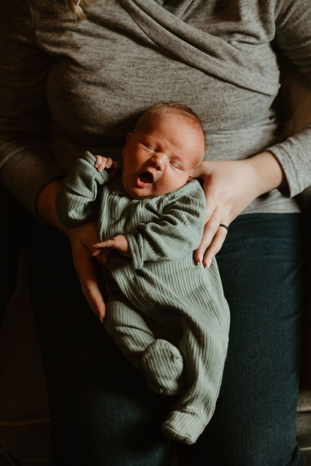 A baby in a sage green outfit yawns while being held against a grey sweater.