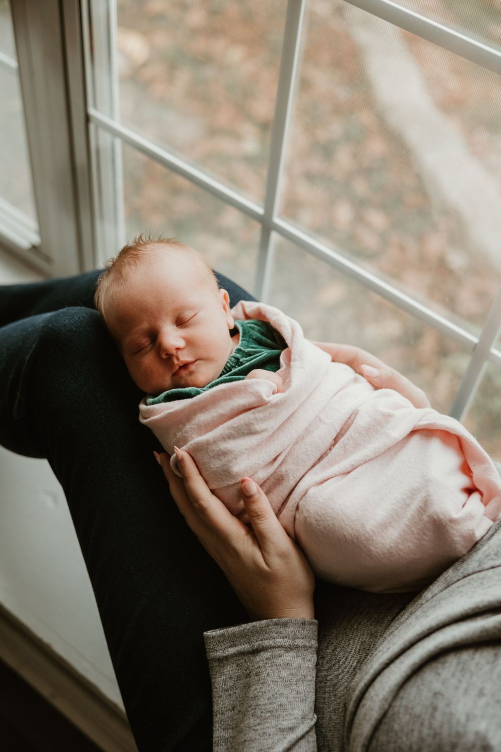 A sleeping newborn wrapped in a pink blanket rests near a window.