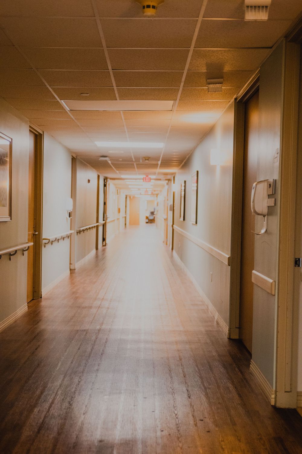 Long empty hospital hallway with fluorescent lighting and wooden doors.