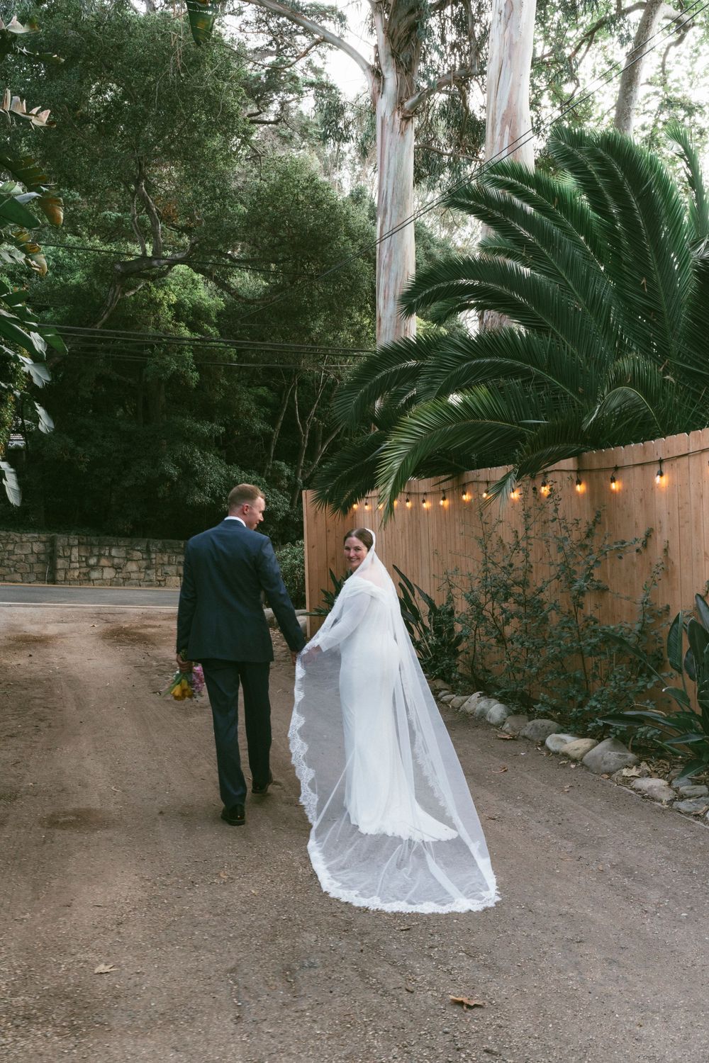 A wedding couple walking down a dirt path surrounded by palm trees and string lights along a stucco wall in Santa Barbara, CA