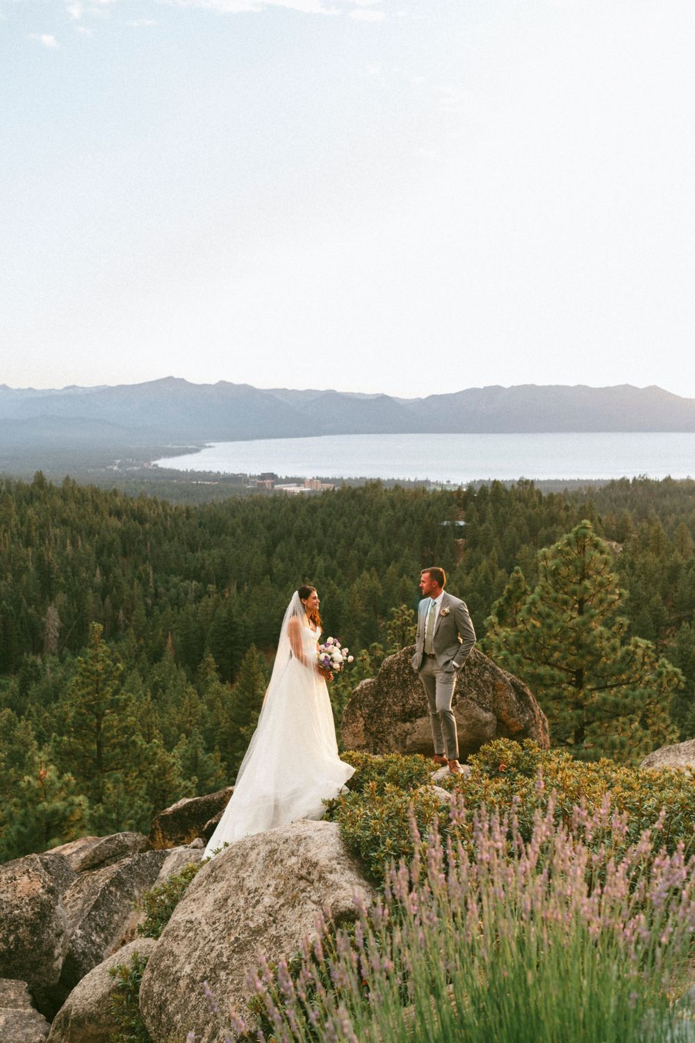 A couple stands on a rocky outcrop overlooking a lake tahoe and mountains during their scenic mountain wedding ceremony.