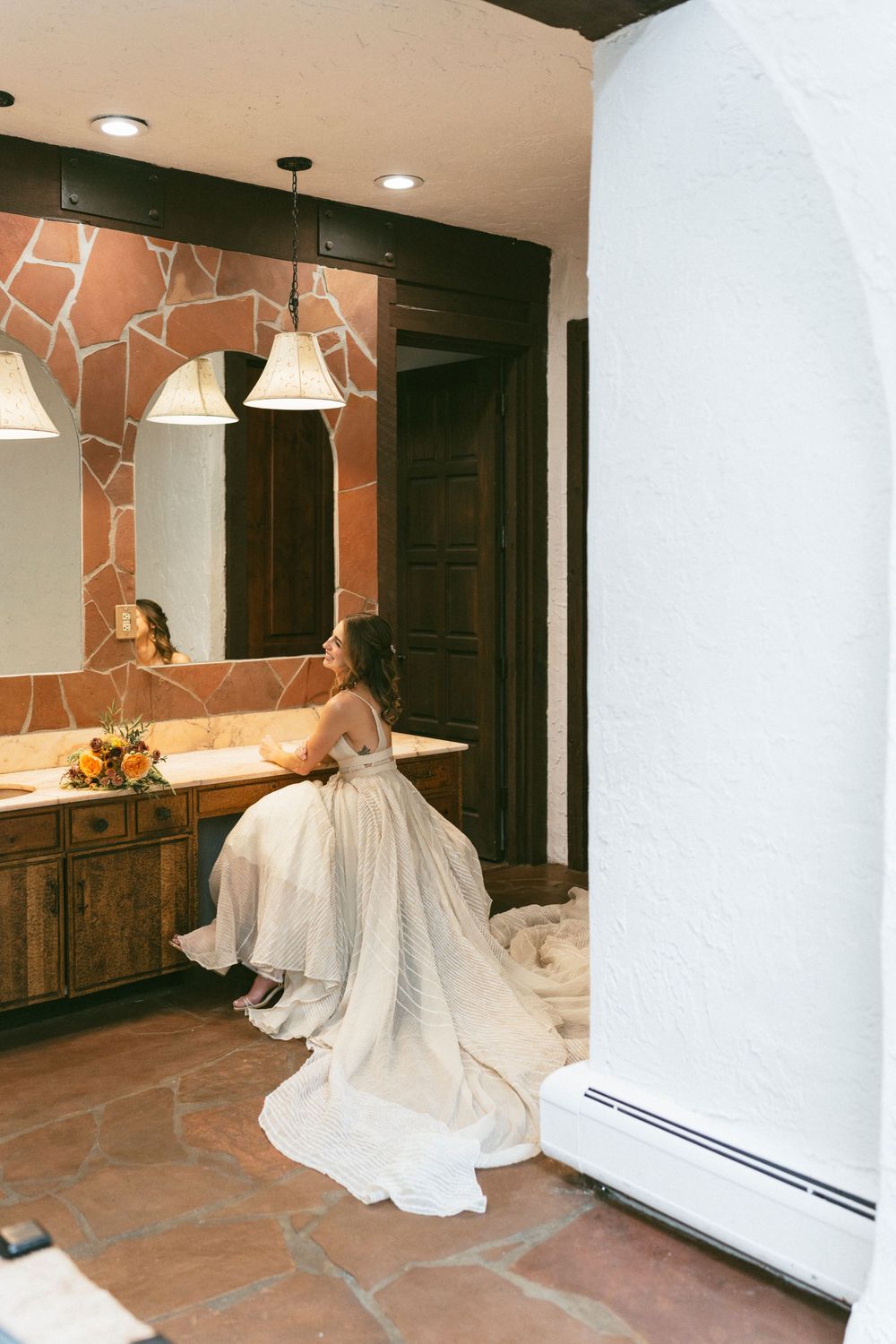 A person in a flowing white wedding gown sits at a rustic bathroom vanity with stone walls and pendant lights at Villa Parker