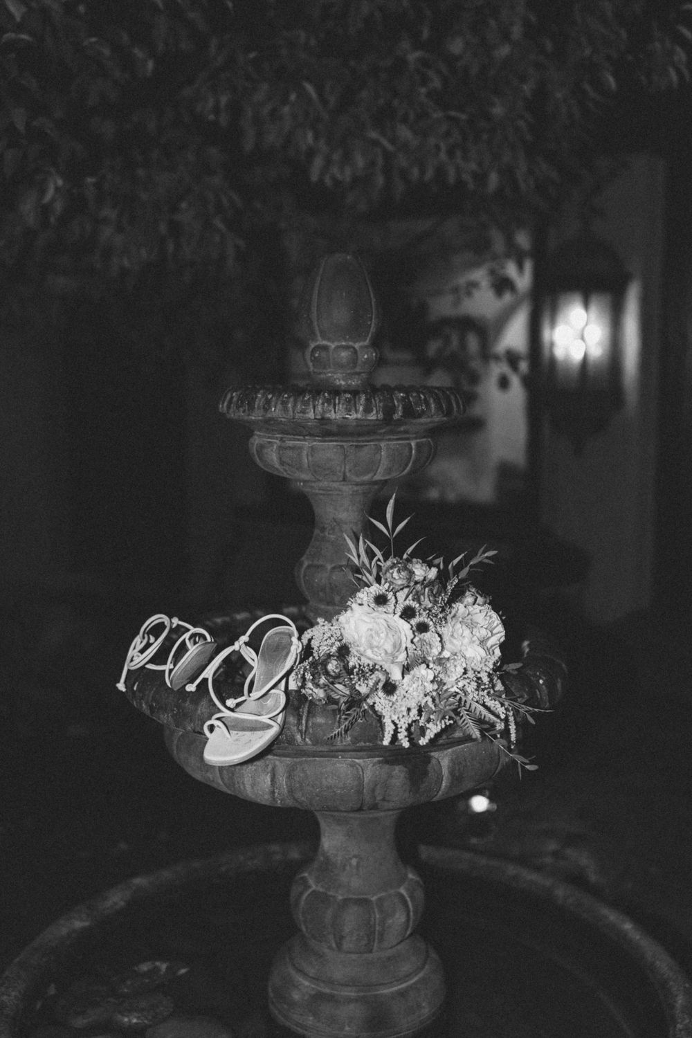 Black and white photograph of bridal shoes and floral arrangement placed on a decorative tiered fountain at Villa Parker in Colorado