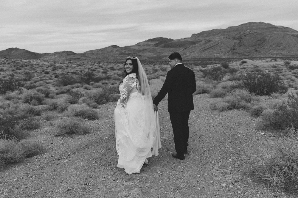 A couple in wedding attire walks along a desert path with mountains in the background in black and white in Las Vegas, Nevada