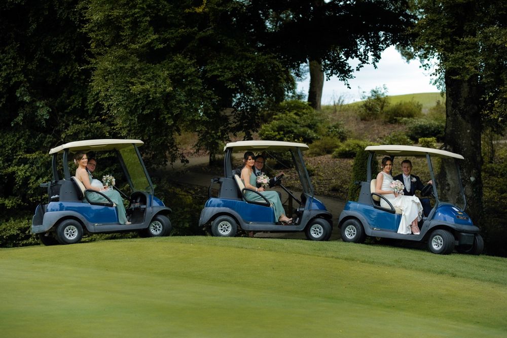 A bride, groom, and wedding party ride in blue golf carts on a lush green golf course, smiling and holding bouquets, New Forest, Tullamore.