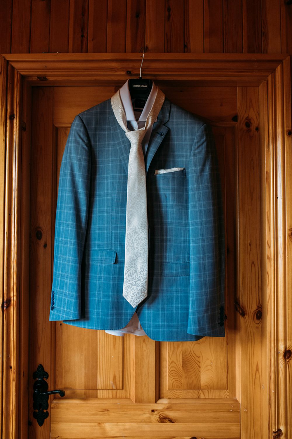 A blue checkered suit jacket with a patterned white tie hangs on a wooden door, ready for a special occasion.