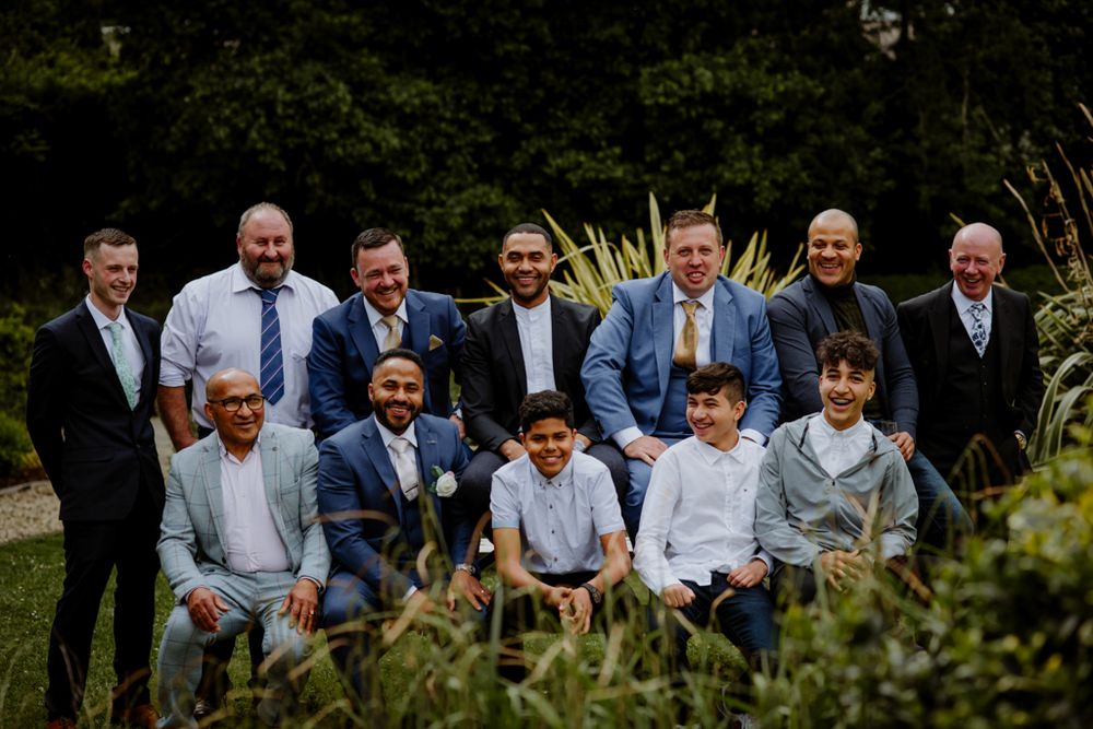 A group of eleven men dressed in formal attire poses together outdoors, surrounded by greenery and decorative plants.