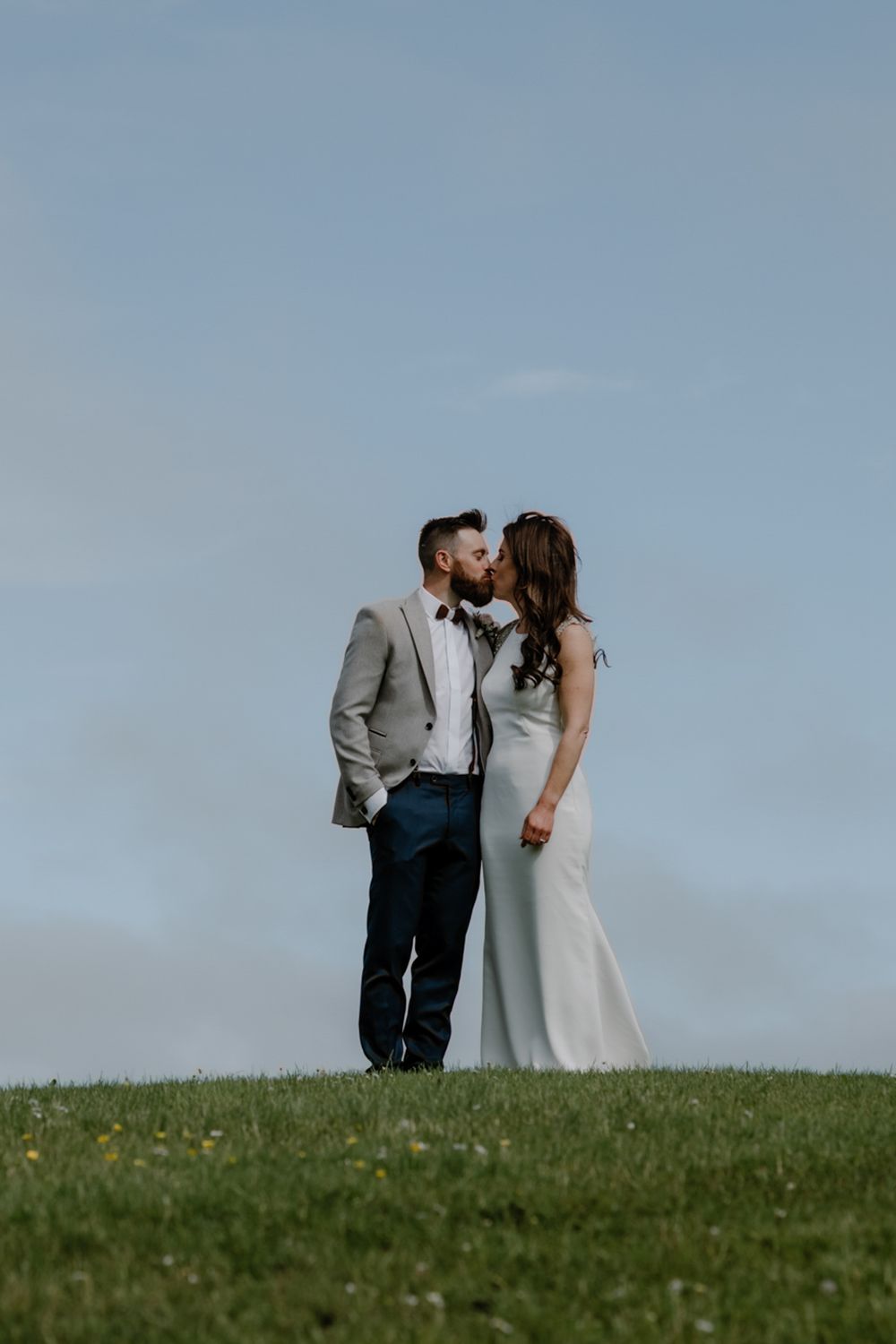 A couple dressed elegantly stands close together on a grassy hill under a clear sky, capturing a romantic moment.