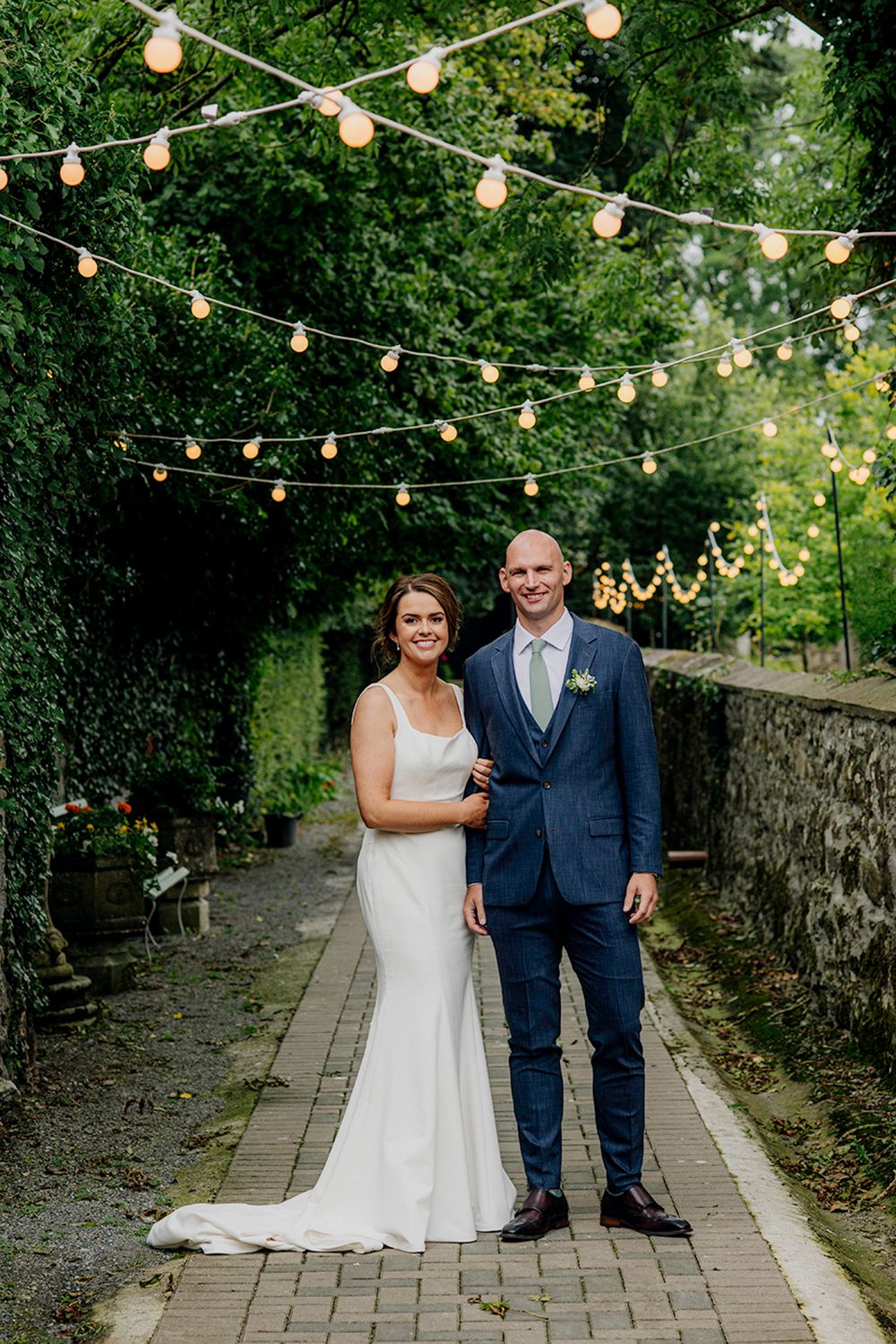A couple stands together in a lush green alleyway, adorned with string lights, joyfully celebrating their wedding day.