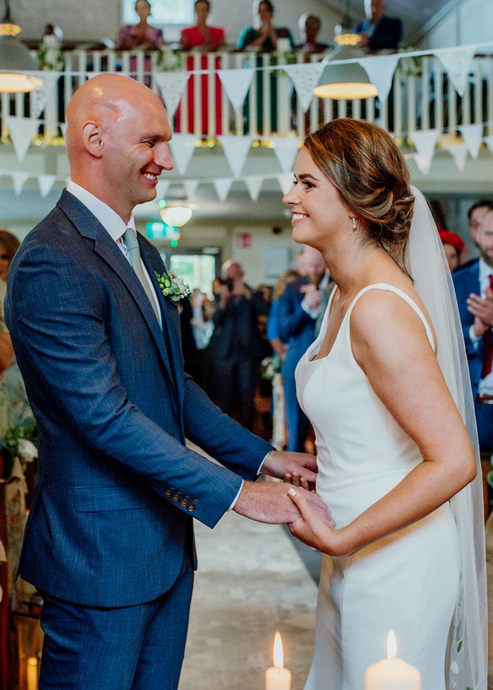 A couple in formal attire stands hand in hand during a wedding ceremony, with guests clapping in a decorated venue filled with candlelight.