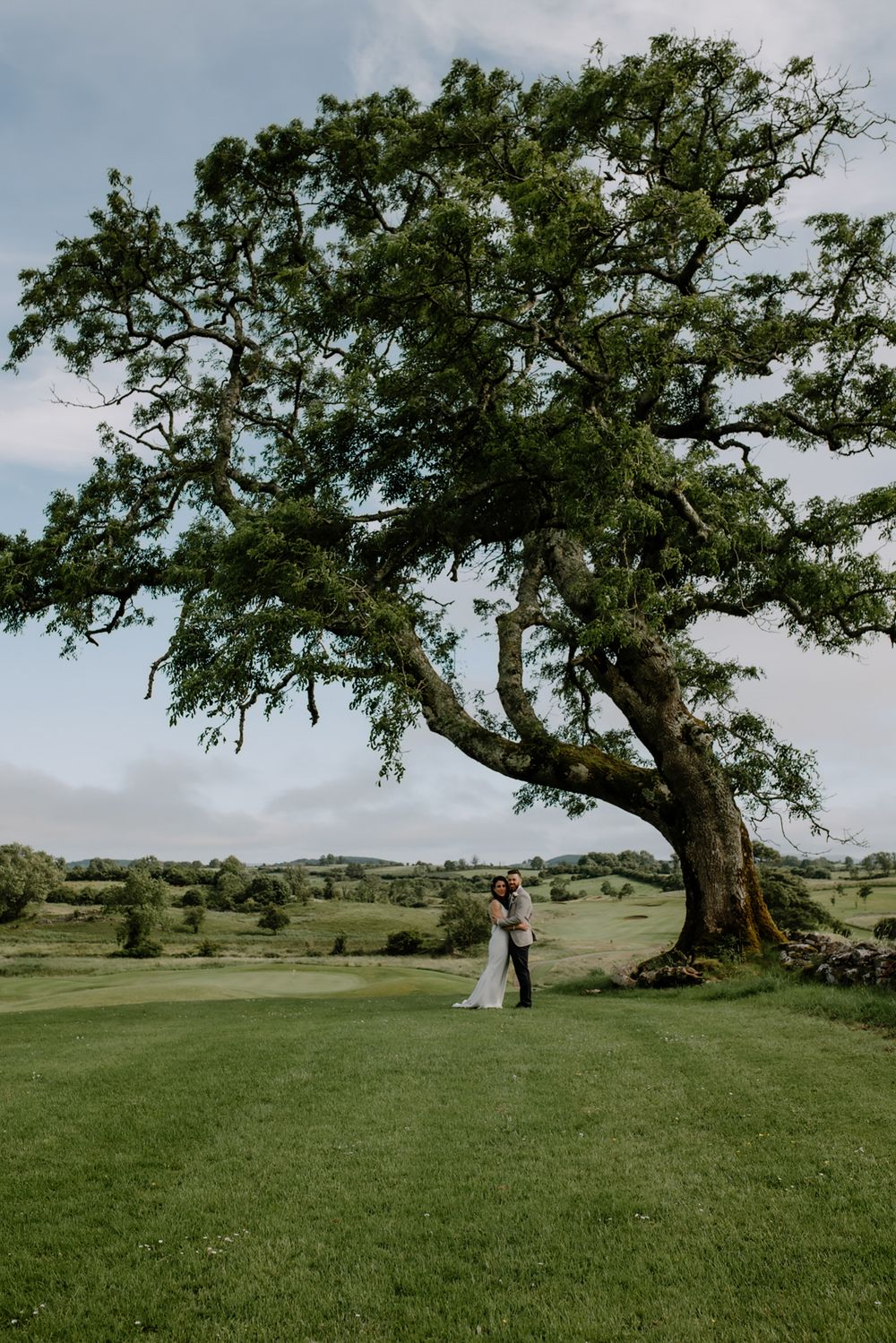 A couple embraces lovingly beneath a large tree in a lush green landscape. Castle Dargan Hotel Sligo.