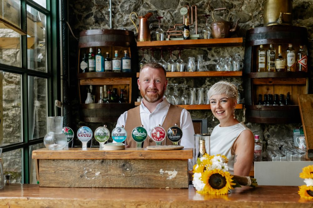 A rustic bar setup featuring beer taps, shelves stocked with spirits, and a bouquet of sunflowers in the foreground. The Barn, Tyrrellspass.