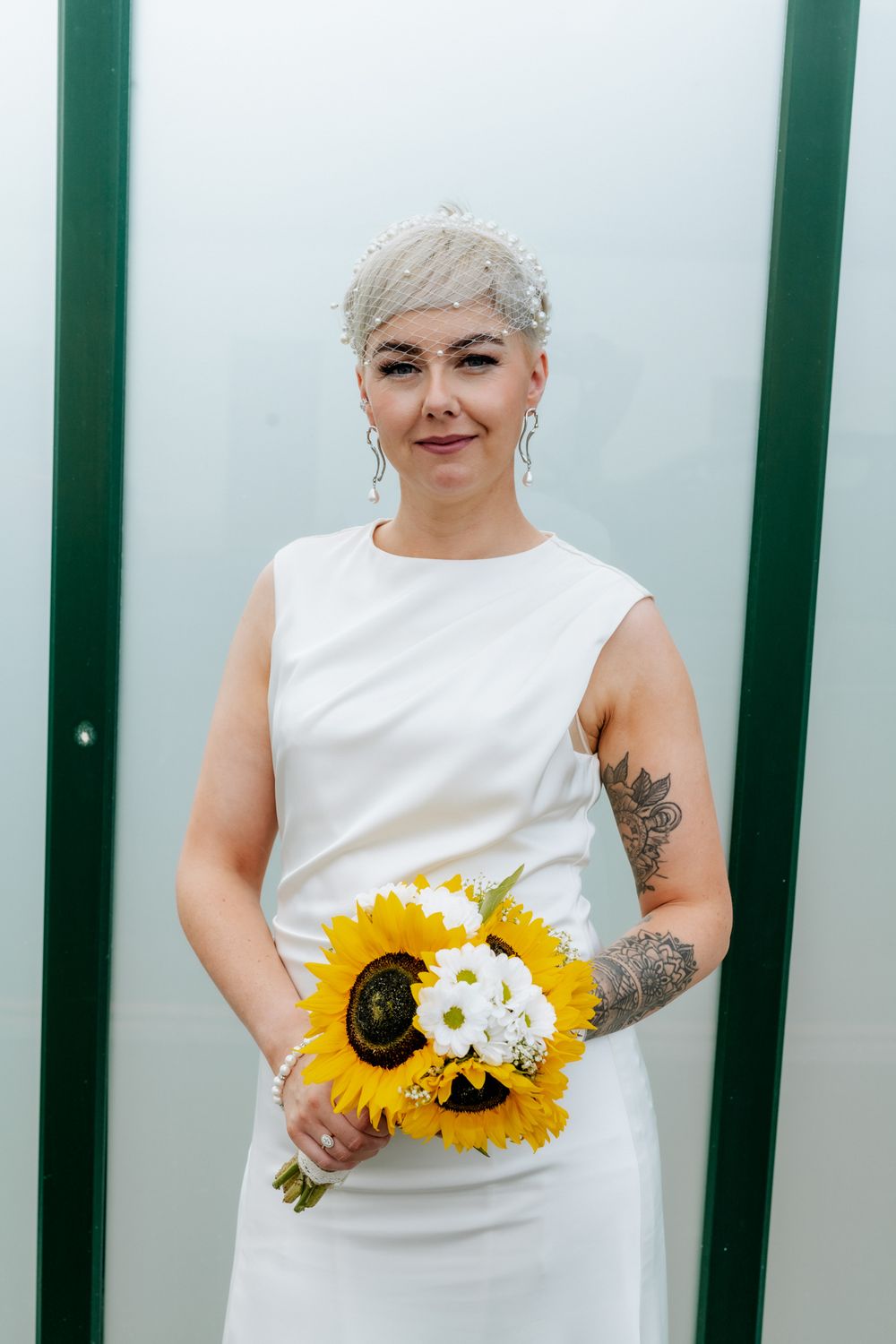 A person wearing a white wedding dress holds a vibrant bouquet of sunflowers and white flowers, with a green background.