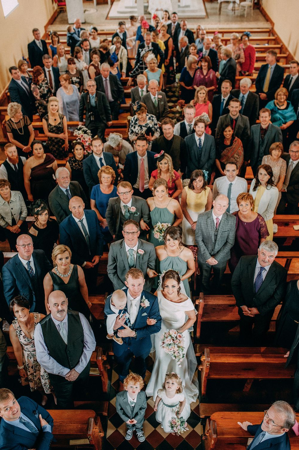 A joyful wedding scene captured from above, showing the bride and groom at the center, surrounded by family and friends in a church.