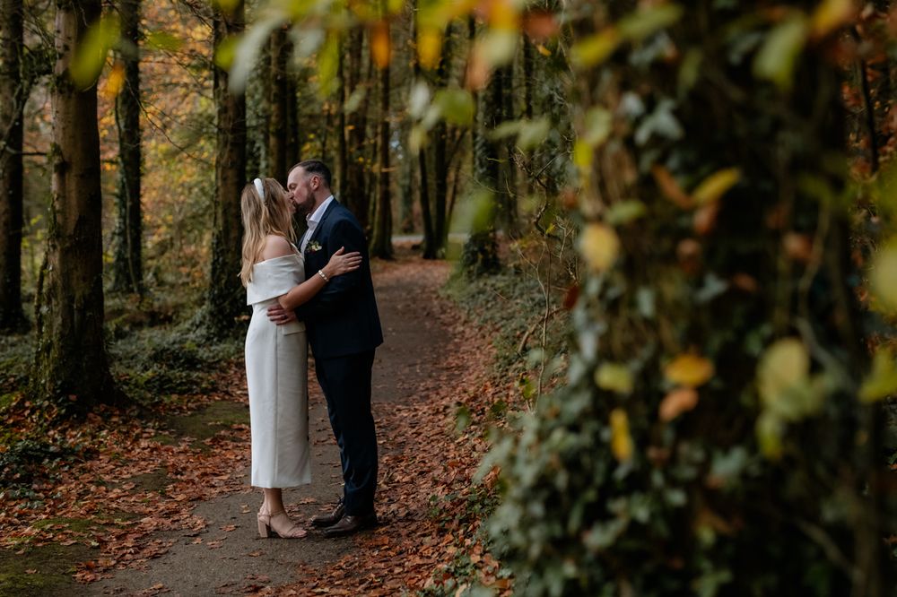 A couple shares a romantic kiss in a forest adorned with autumn leaves, surrounded by tall trees and a peaceful path. Mote Park, Roscommon.
