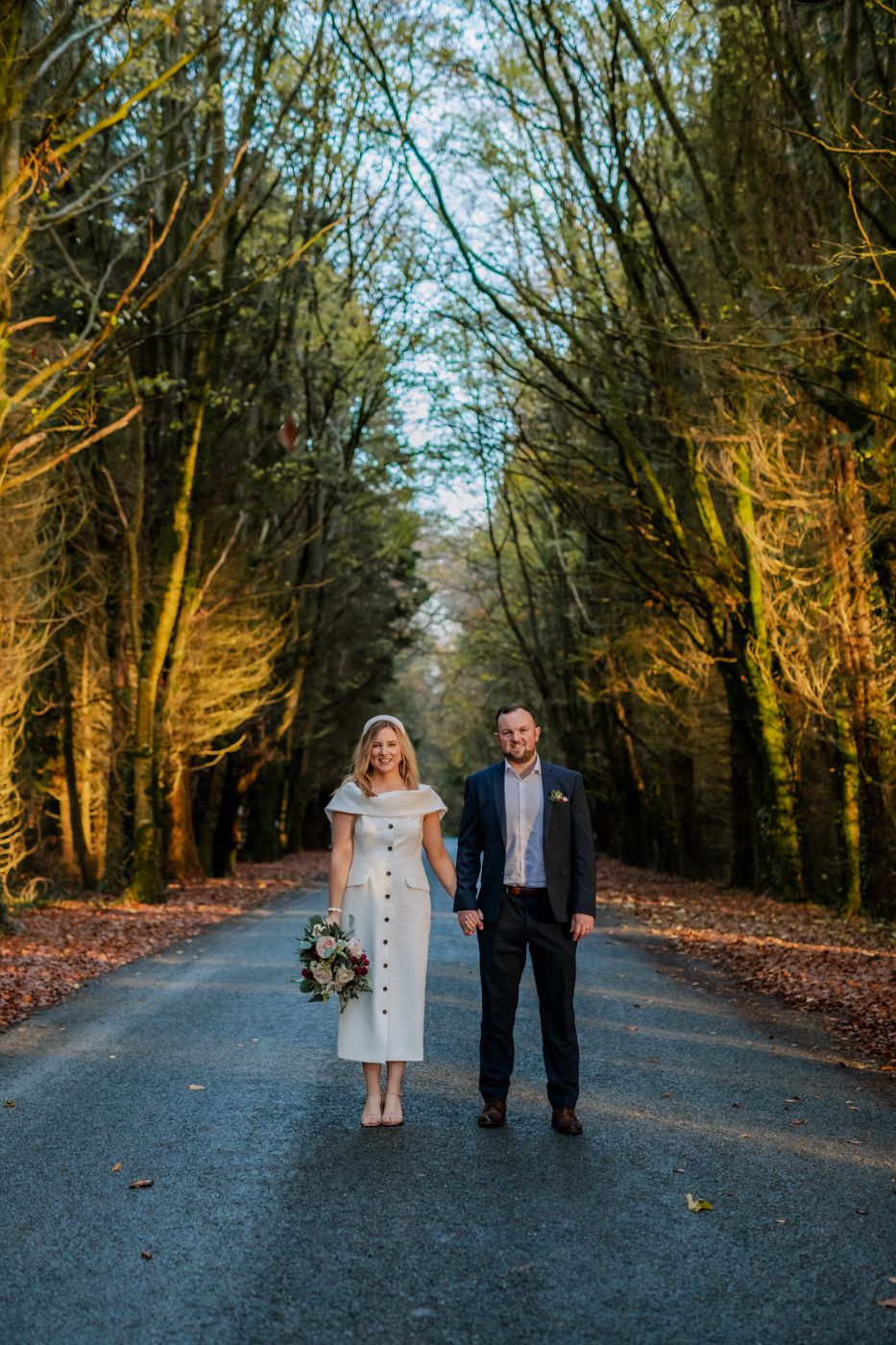 A couple stands hand in hand on a wooded road, surrounded by tall trees and autumn leaves, with the sunlight filtering through branches.