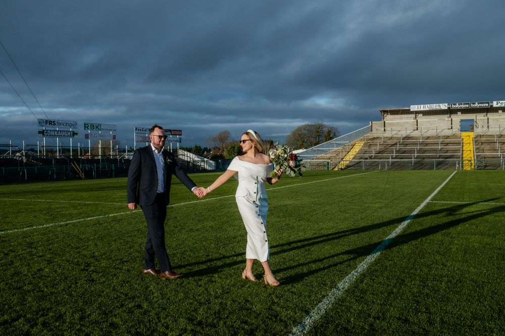 A couple walks hand in hand on a grassy field, the woman in a white dress holding a bouquet, with stadium seating in the background.