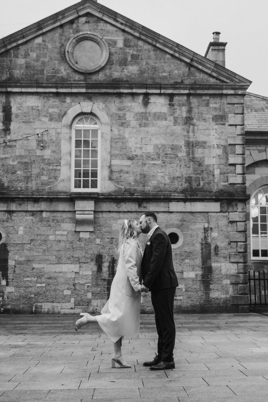 A romantic black-and-white photograph of a newlywed couple sharing a sweet kiss in front of a historic stone building in Roscommon.