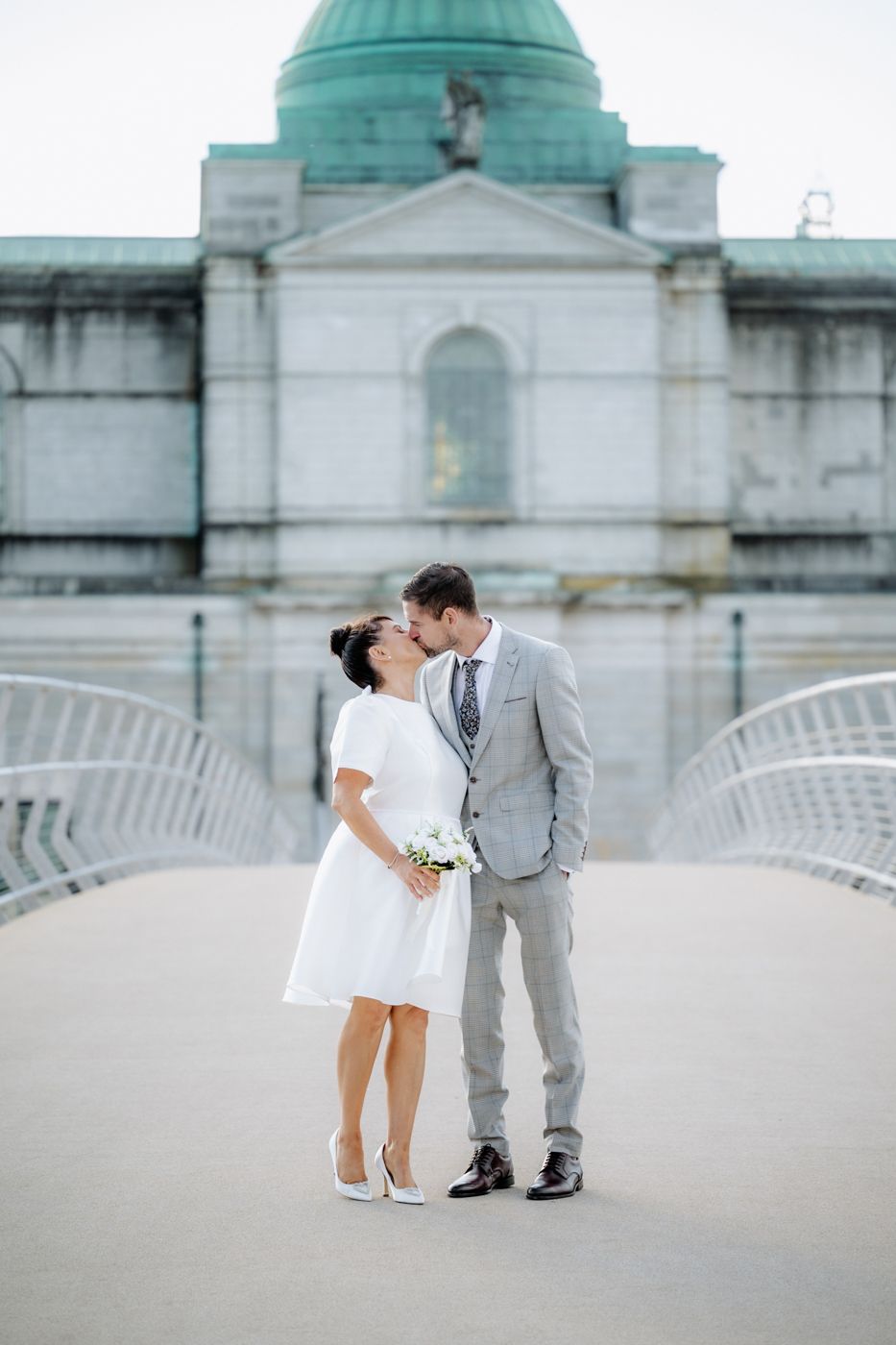 A couple in elegant wedding attire embraces on a bridge, with a historic building and dome in the background, creating a romantic scene.