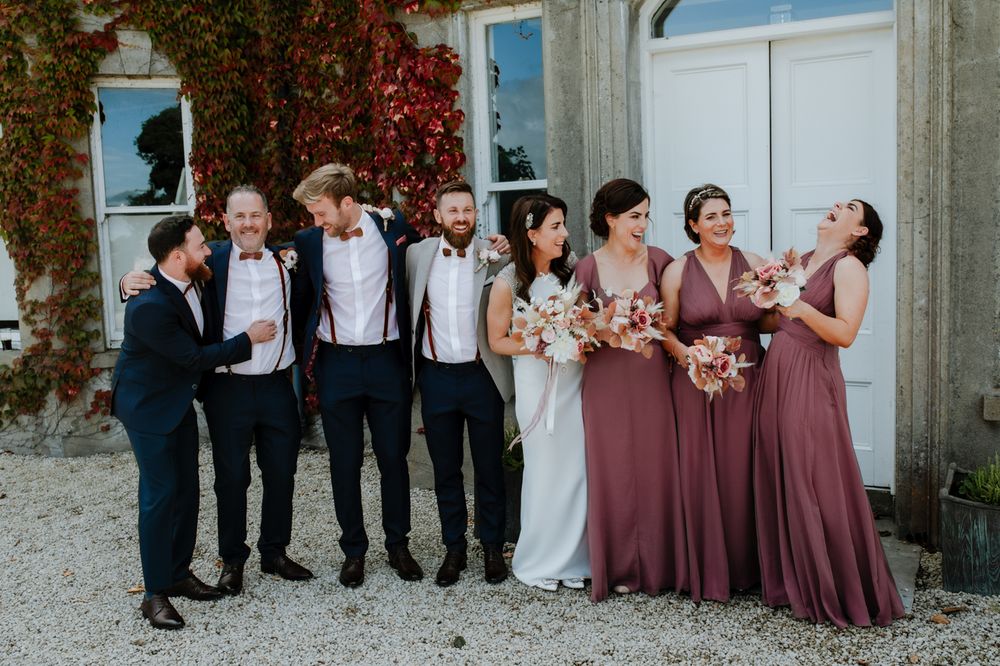 A joyful wedding party poses together outdoors, dressed in elegant attire with flowers, against a backdrop of vibrant ivy-covered walls.
