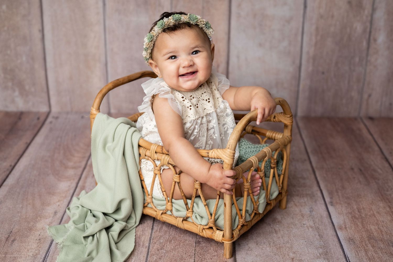 Small child in white dress sits next to vintage wooden doll cradle on rustic wooden floor.