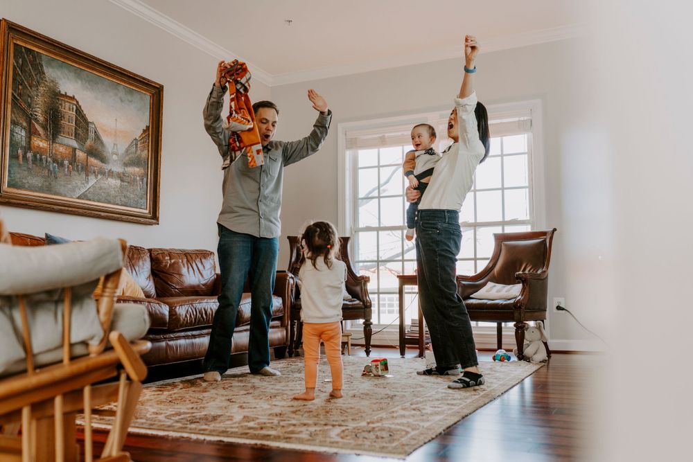 Fun family photography of a whole family dancing and jumping with joy together in-home in their bright living room.