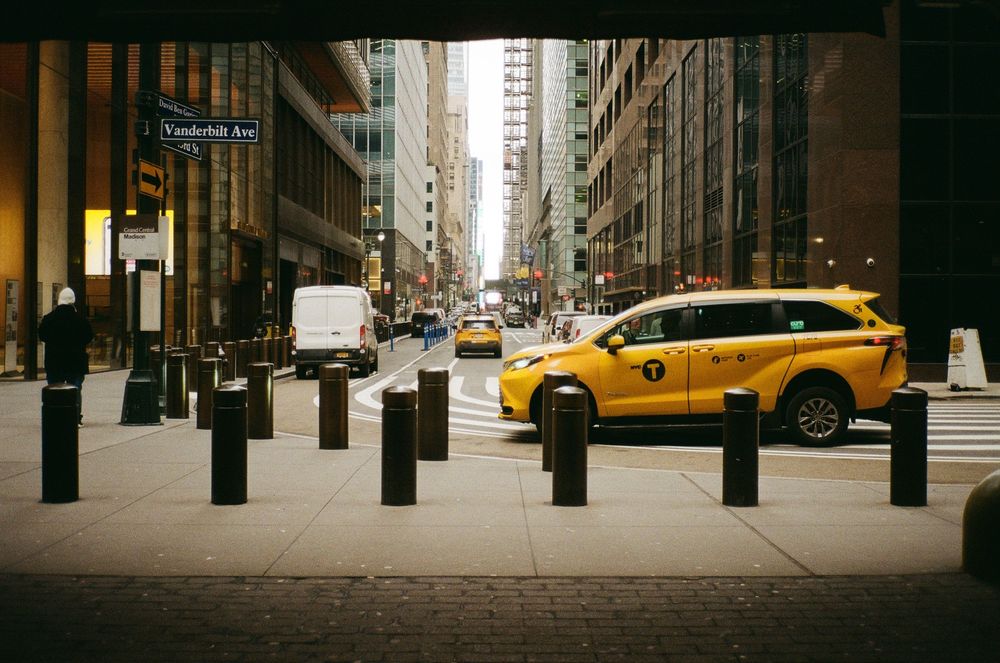 An iconic yellow taxi drives past modern high-rise buildings on a busy street in downtown Manhattan.